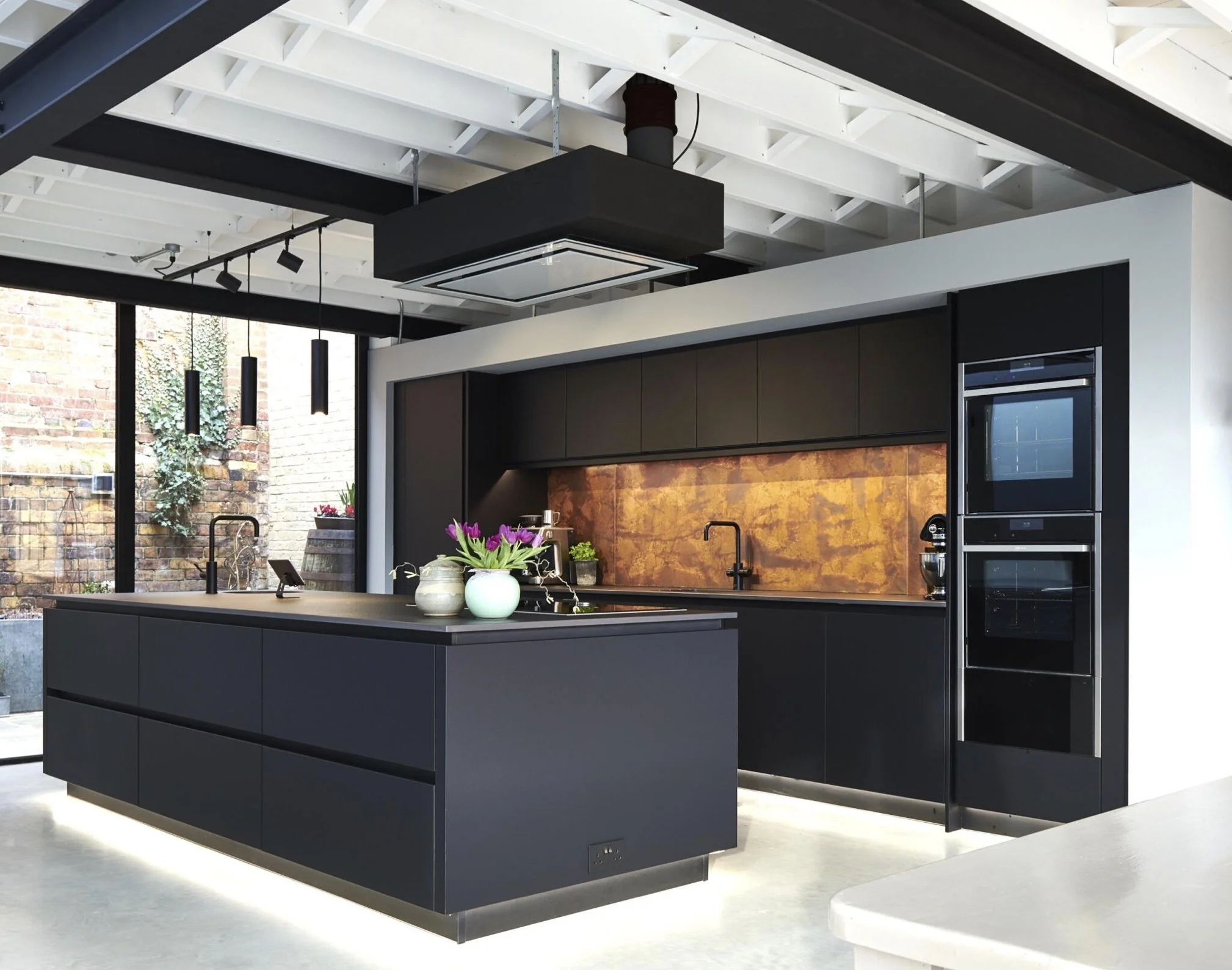 Modern black kitchen with island, built-in oven, and black cabinets, decorated with flowers and plants, with large windows showing an outdoor brick wall.