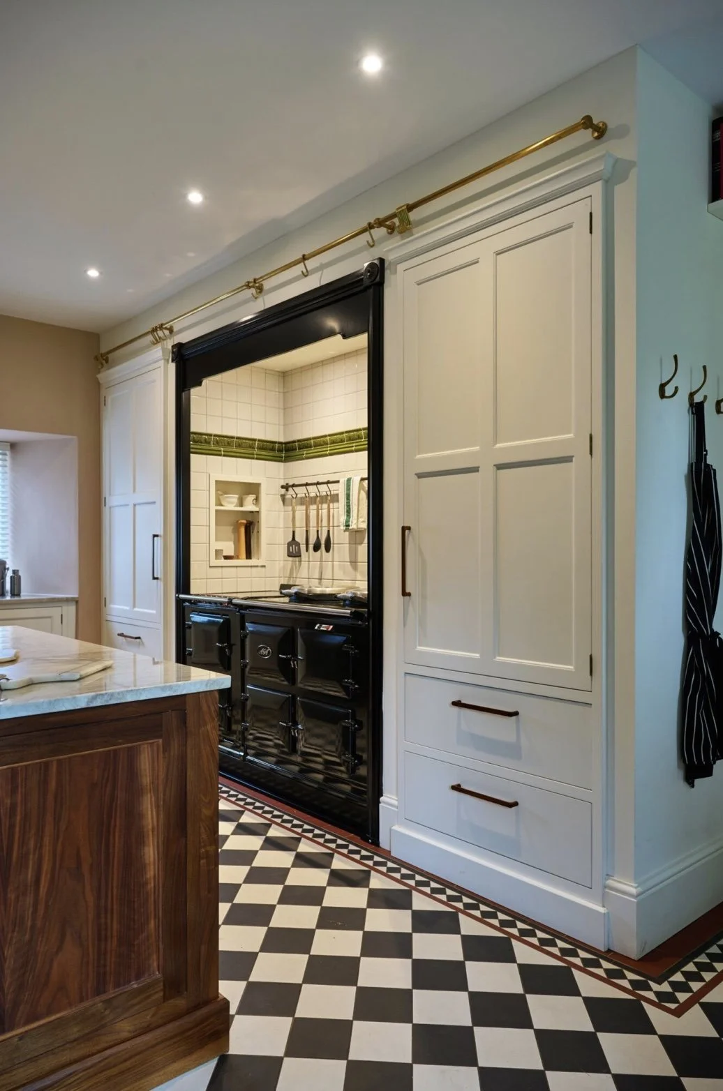 A kitchen with a vintage black stove in a small alcove, white tiled walls with green accents, white cabinetry, a wooden island, and checkered black and white floor tiles.