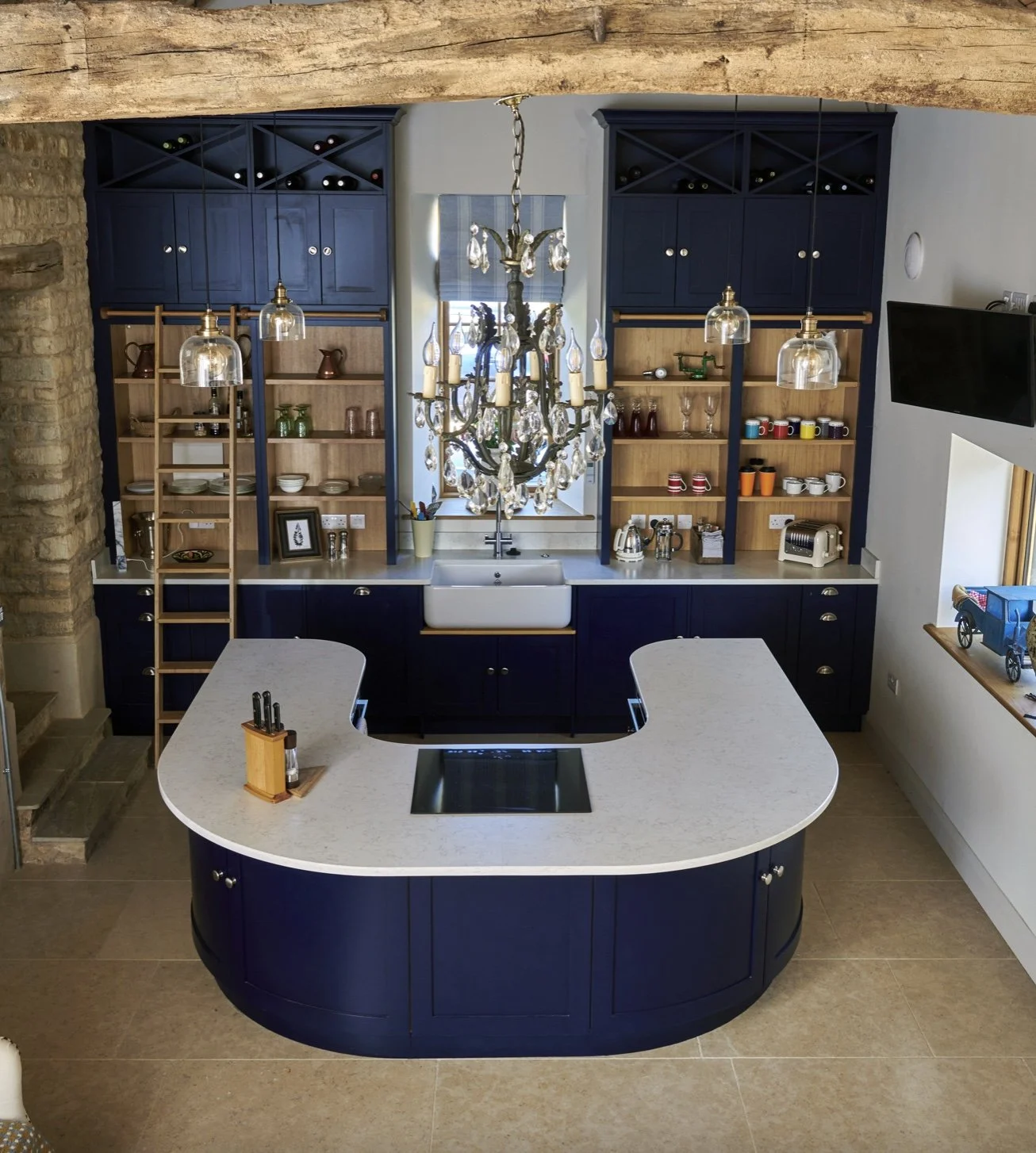 A modern kitchen with dark blue cabinets, open wooden shelves, and a white dual-level countertop island. A chandelier hangs above the sink, and there is a window with a shade behind the sink. Appliances and decorative items are on the shelves and cou