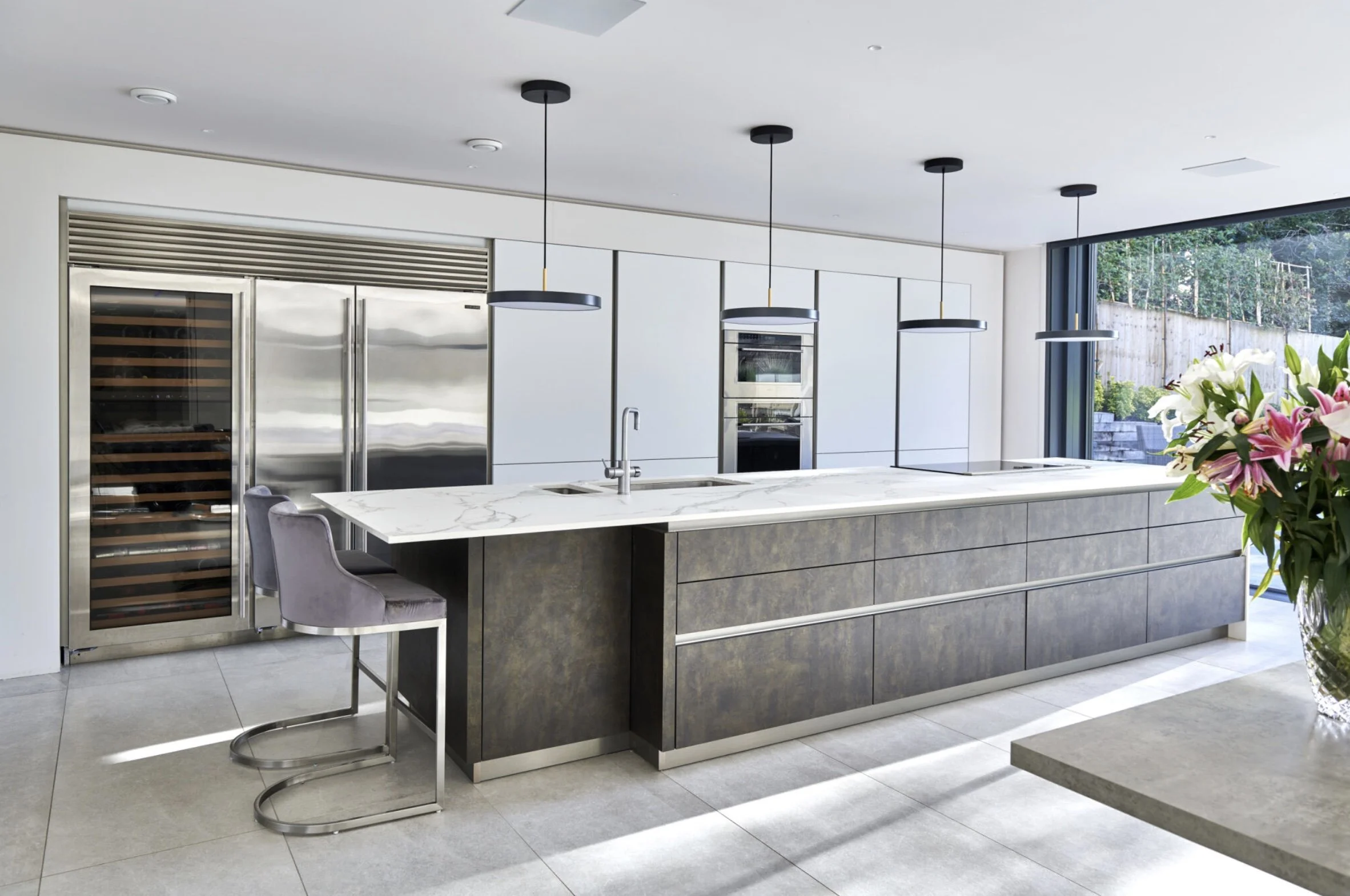 Modern kitchen with large island, white marble countertop, gray cabinetry, stainless steel appliances including a wine cooler, and black pendant lights hanging above the island. Large window with outdoor view and a vase of pink and white flowers on a