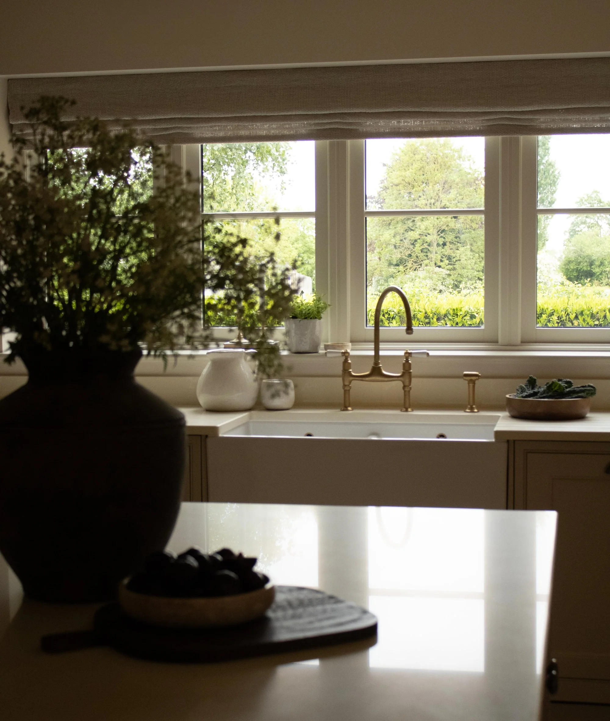 Kitchen window with view of trees and greenery outside, gold-colored faucet at the sink, and various potted plants and decor on the windowsill, with a floral arrangement in the foreground.