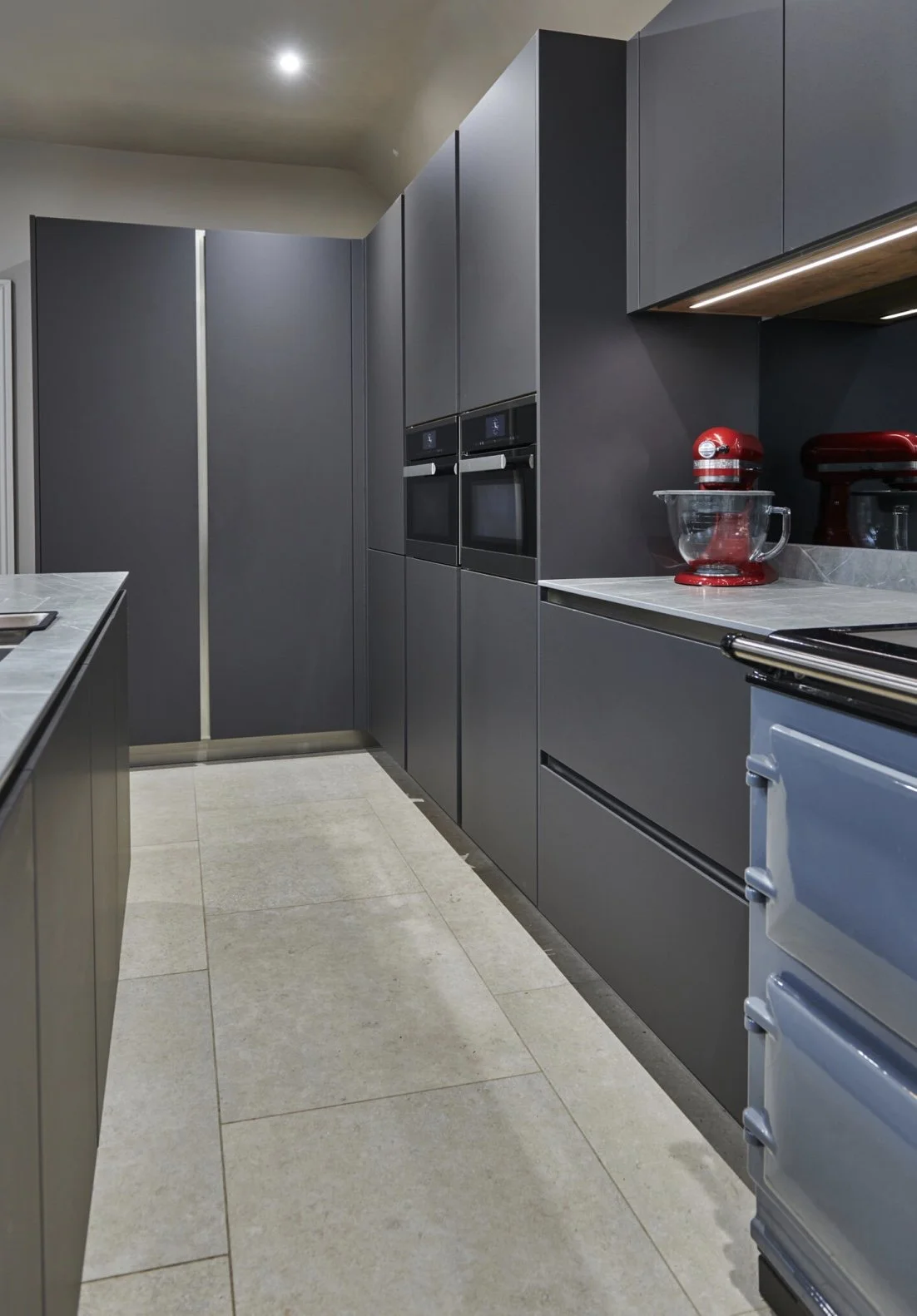 Modern kitchen with dark gray cabinets, built-in ovens, a gray countertop, and red kitchen appliances including a stand mixer and a blender. The floor is tiled in light beige.