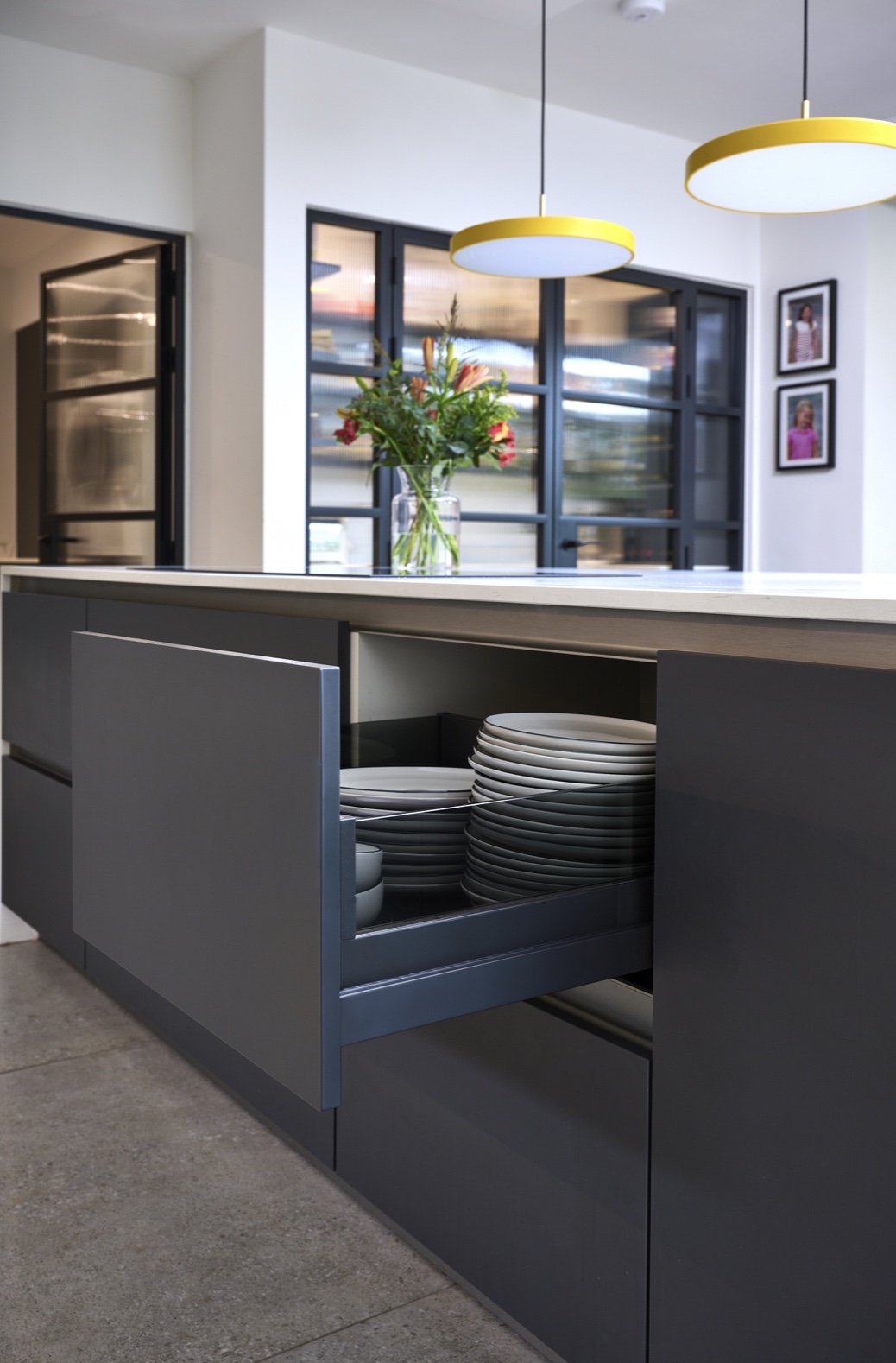 Open kitchen drawer filled with stacked white plates with a modern, minimalist design. In the background, there is a white countertop with a vase of colorful flowers, a window with black framing, and two hanging yellow-and-white pendant lights.