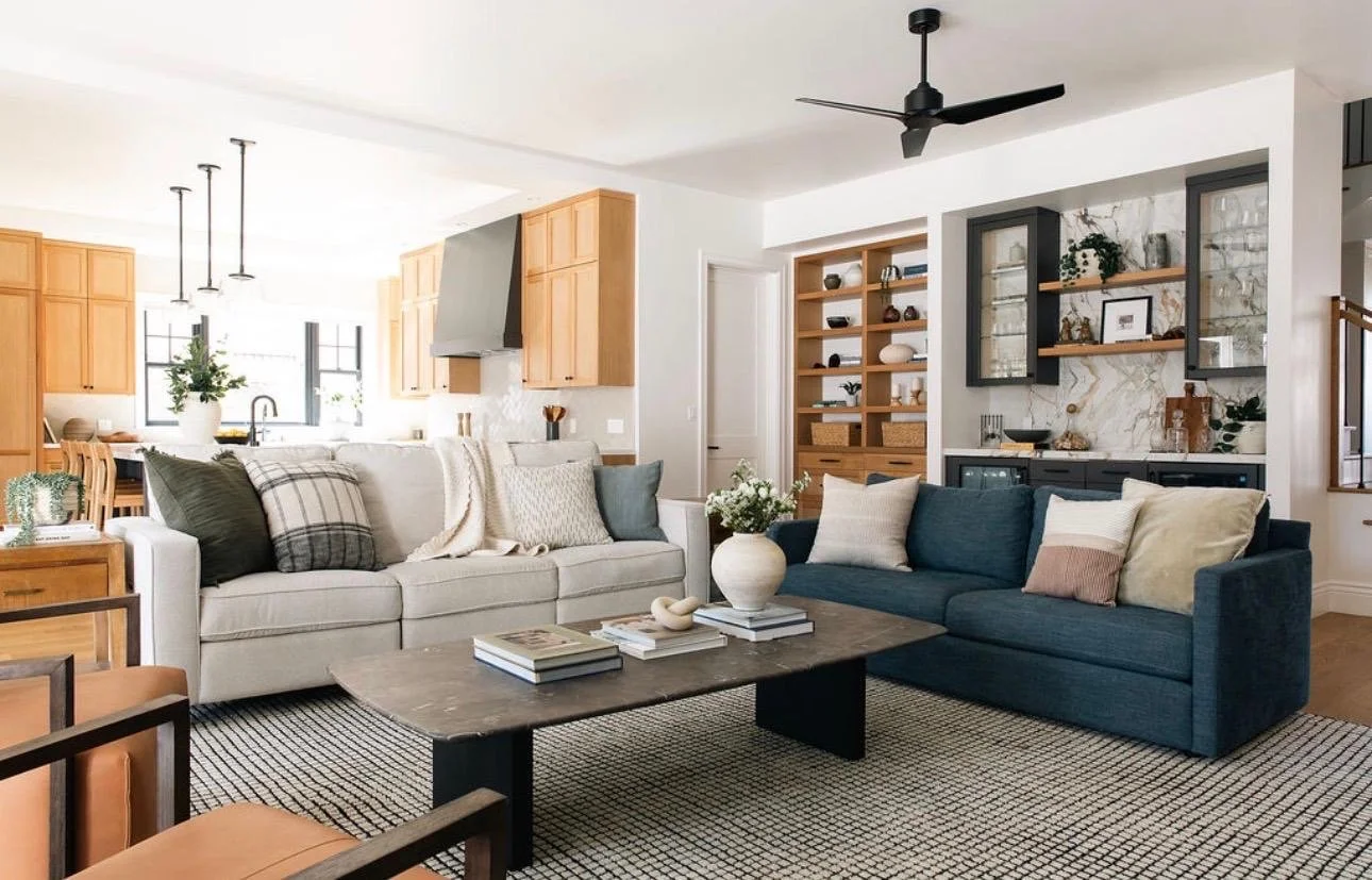 Modern living room with a navy blue sofa, white sofa, wooden coffee table, and decorative pillows. Open kitchen visible in background with wooden cabinets, pendant lights, and a large window. Books and decorative items on shelves and a patterned rug 