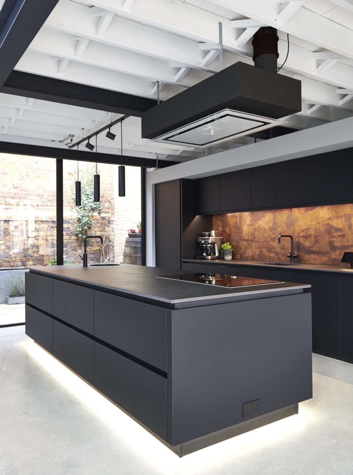 Modern kitchen with black cabinetry, a large island, built-in stovetop, and pendant lights, with an industrial ceiling and large window.