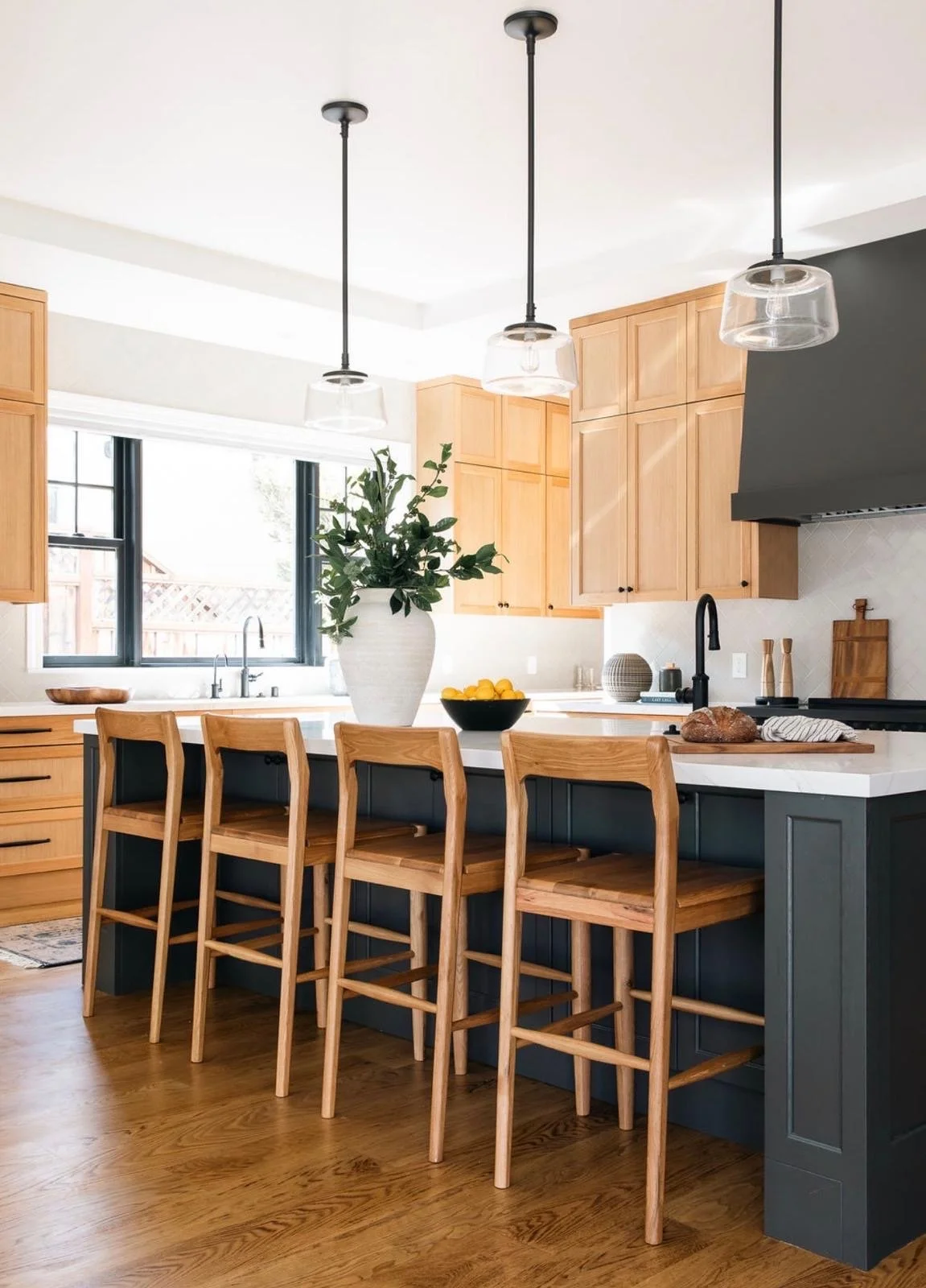 Modern kitchen with wooden cabinets, a white countertop, black farm sink, and a dark blue island with four wooden chairs. Decor includes a large white vase with greenery, a bowl of lemons, and various kitchen items, all under three hanging pendant li