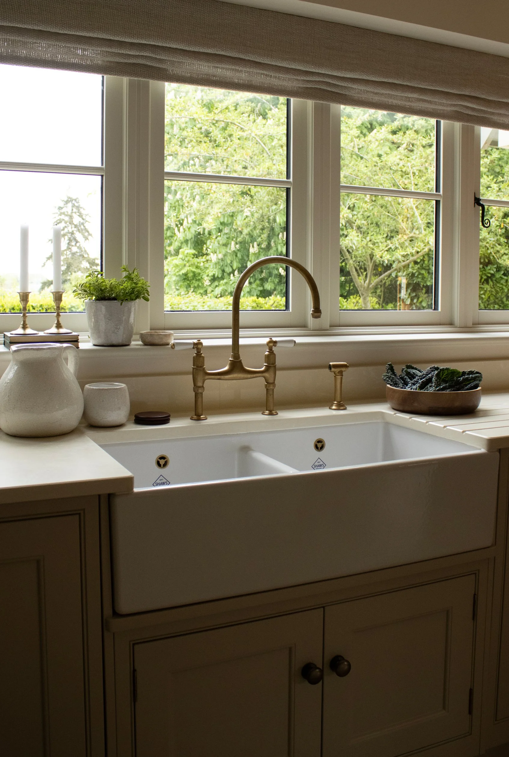 Kitchen window with beige curtains, view of green trees outside, countertop with potted plant, two lit candles, a pitcher, bowls, and a black dishcloth.