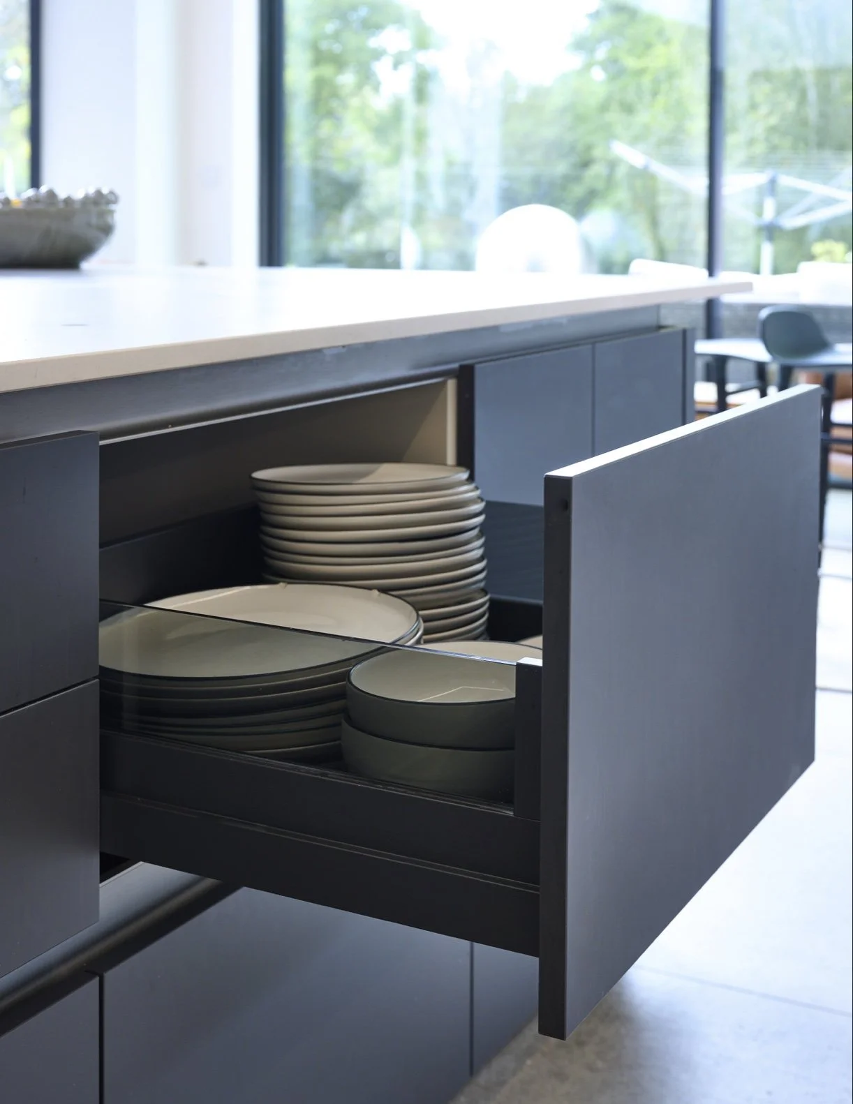 Kitchen cabinet with open drawer containing stacked white and gray plates, with a modern, minimalist design and a view of a bright, outdoor patio through large glass windows.