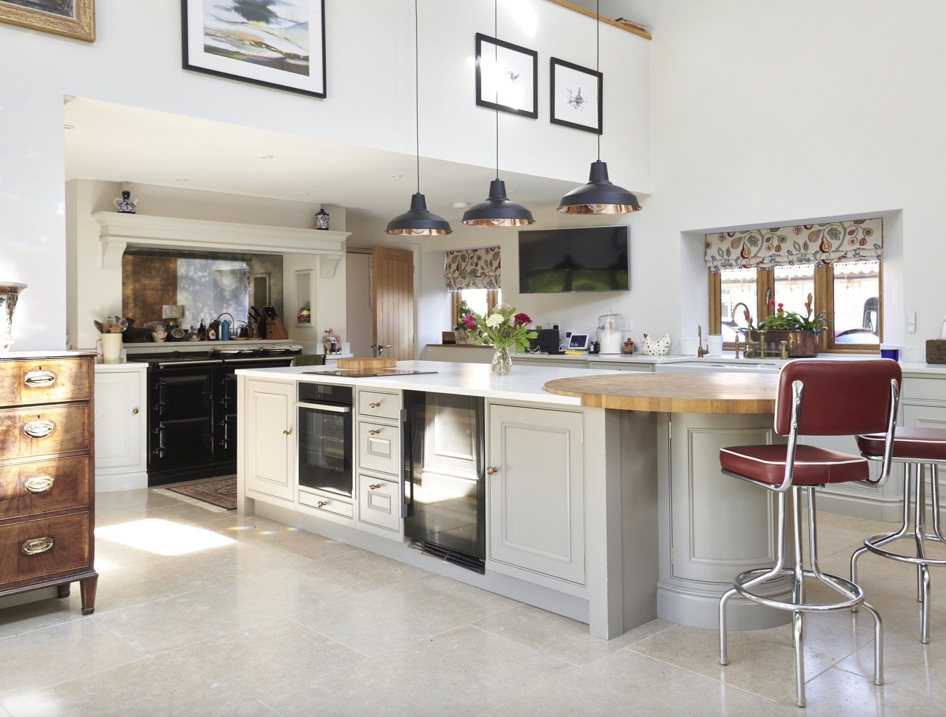 A bright open-concept kitchen with a large island, white cabinets, and a built-in wine cooler, featuring three black pendant lights, a flat-screen TV, and two red barstools.