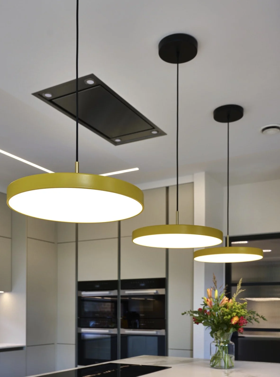 Modern kitchen with three yellow pendant lights hanging above a white kitchen island, a vase of colorful flowers on the island, and a built-in oven and microwave in the background.