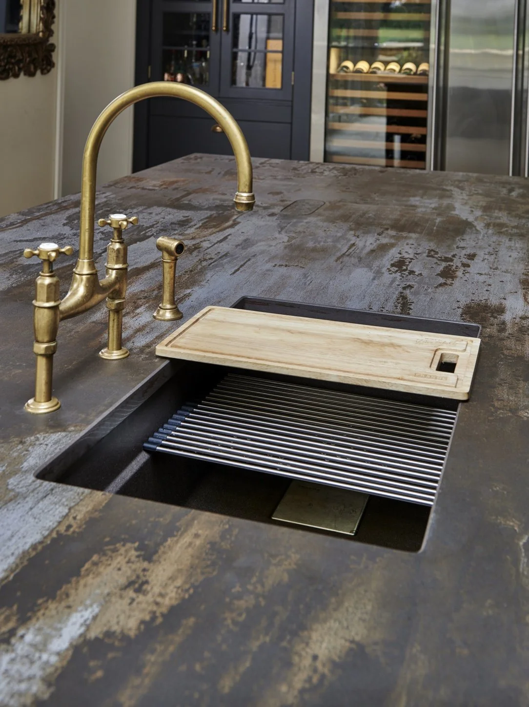 A worn kitchen or bar counter with a bronze faucet and sink, a wooden cutting board, and a rack with metal rods, with a wine fridge and cabinet in the background.