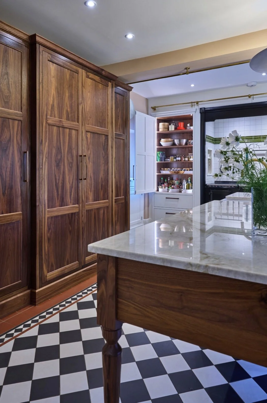 A kitchen with wooden cabinets, a marble countertop, and a black and white checkered floor, with an open pantry and a window with a curtain rod.