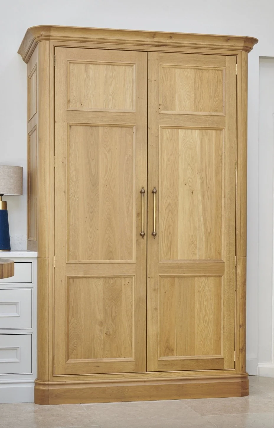 A tall wooden wardrobe with double doors and metal handles, standing next to a white dresser with drawers and a table lamp on top.