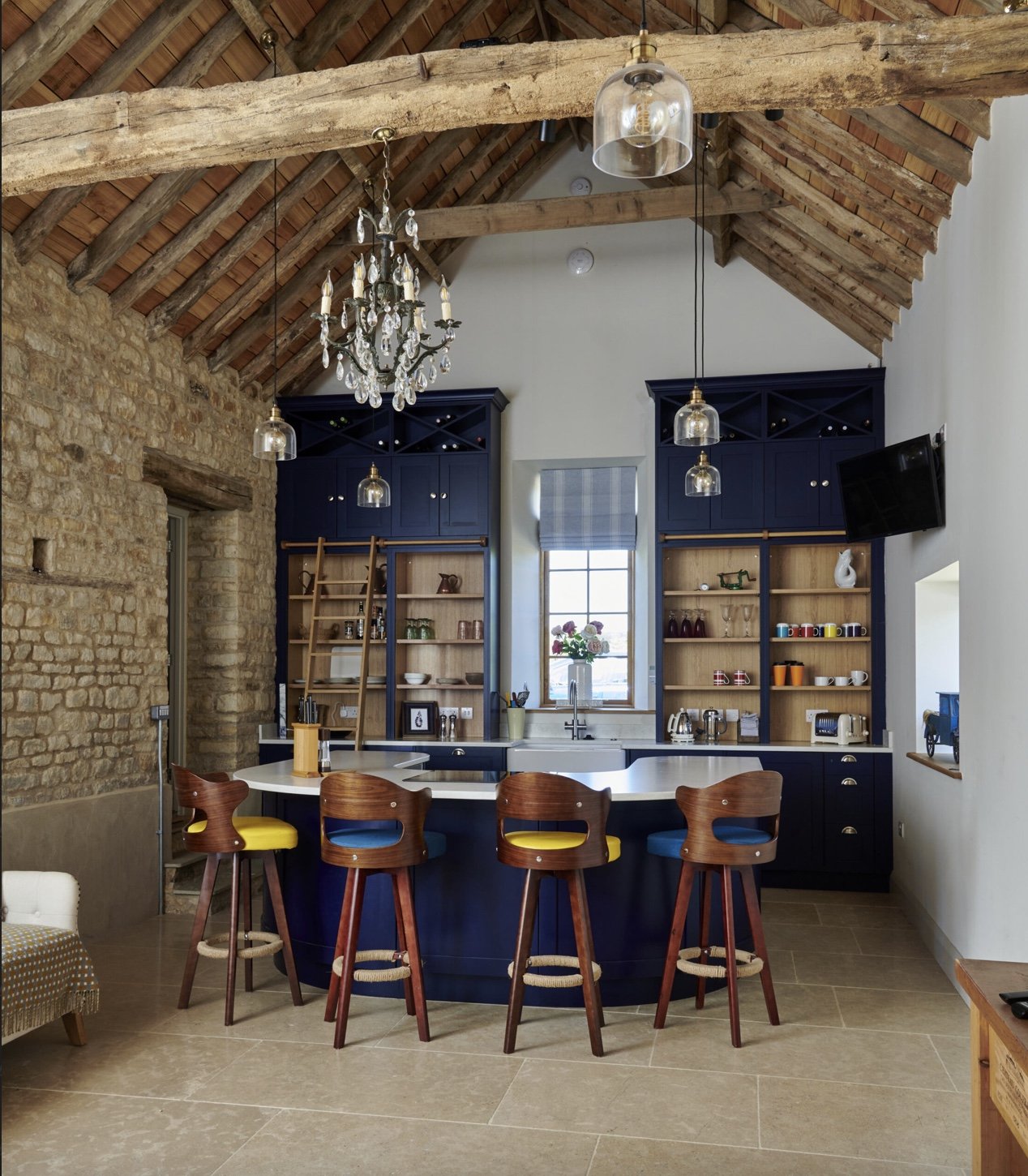 Kitchen with blue cabinets, a central curved island with colorful bar stools, open shelving, stone wall, chandelier, and hanging lights.