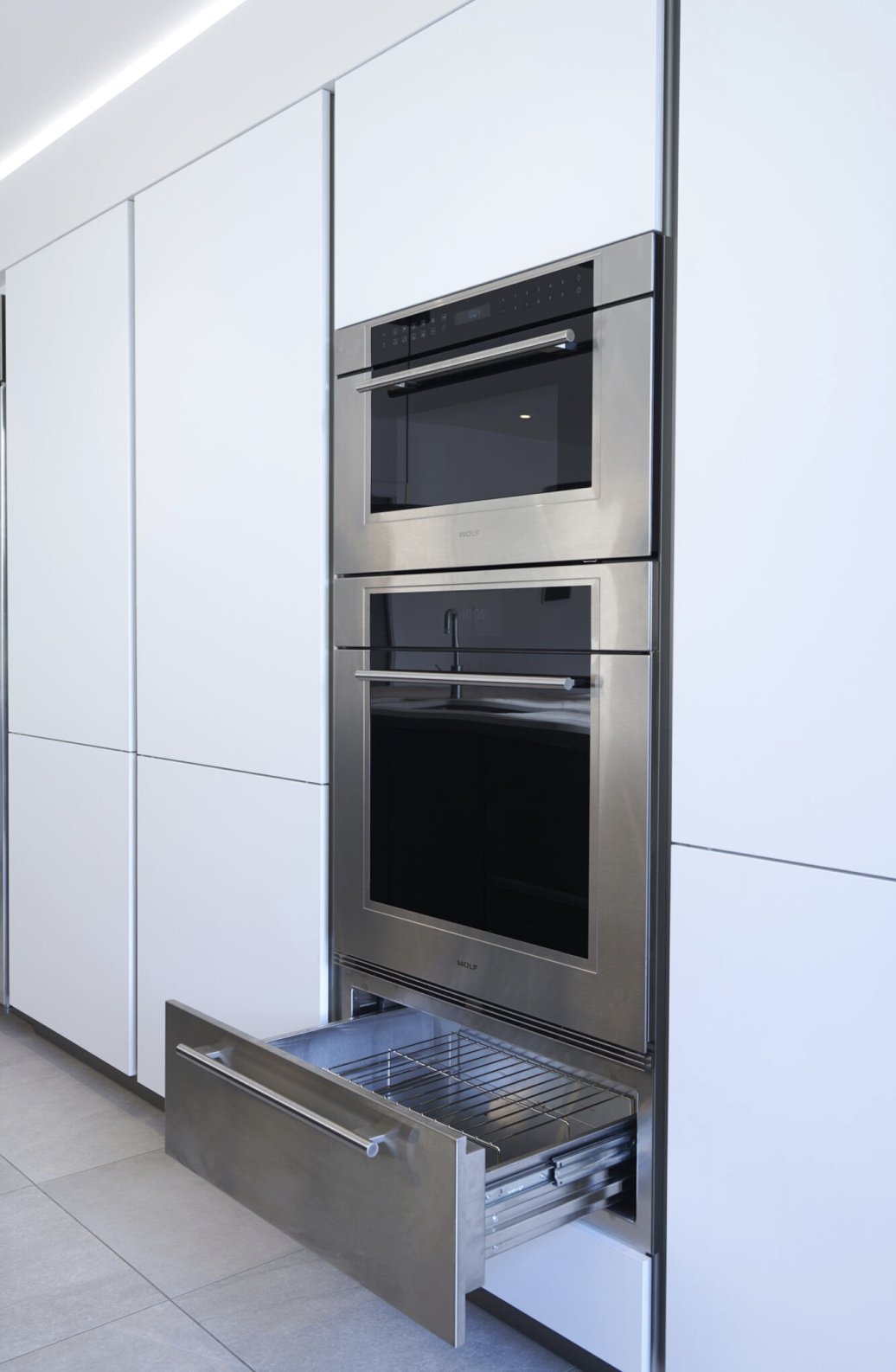 Modern kitchen with built-in stainless steel oven and microwave, white cabinetry, and a partially pulled out metal drawer.