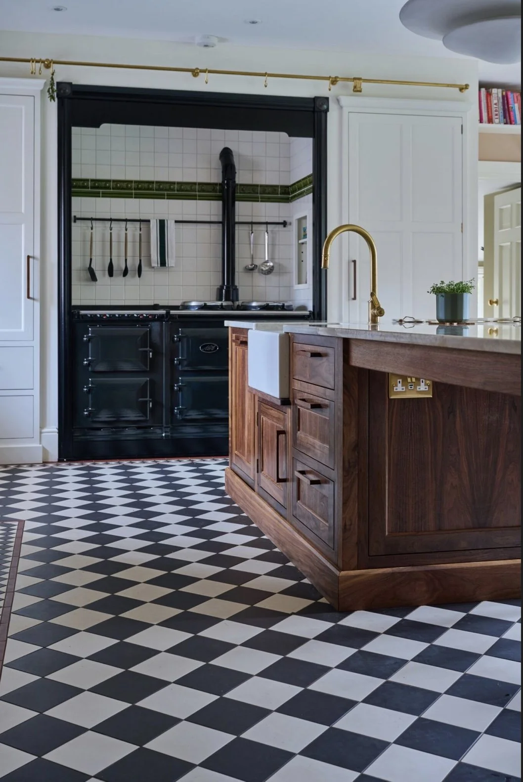 View of a kitchen with a black wood-fired oven and stove behind a slide-open window, a wooden kitchen island with a sink and a gold faucet, black and white checkered floor tiles, and white cabinetry.