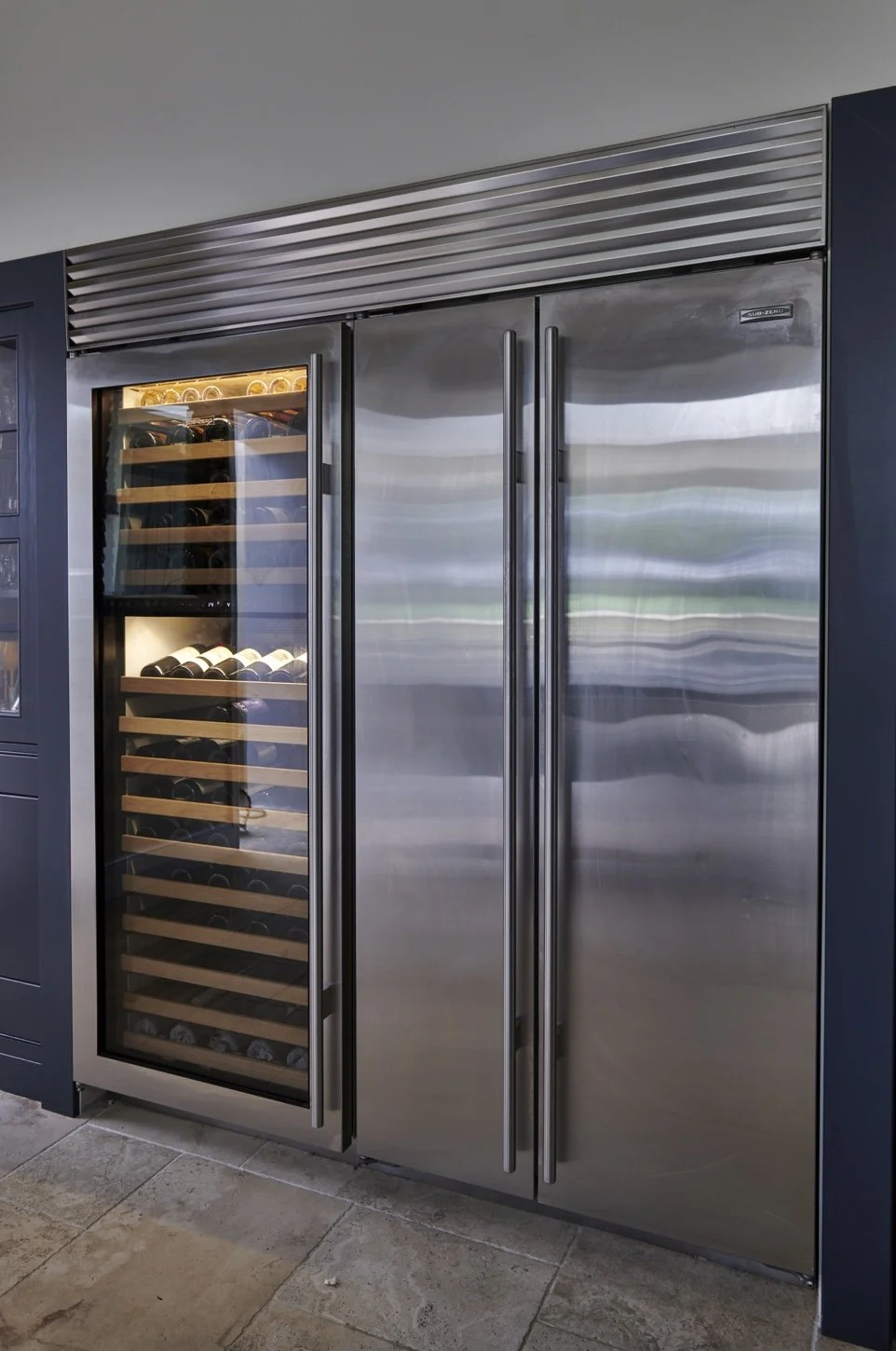 Stainless steel wine refrigerator with a glass door, containing multiple wine bottles stored on wooden shelves, located beside a silver refrigerator with double doors.