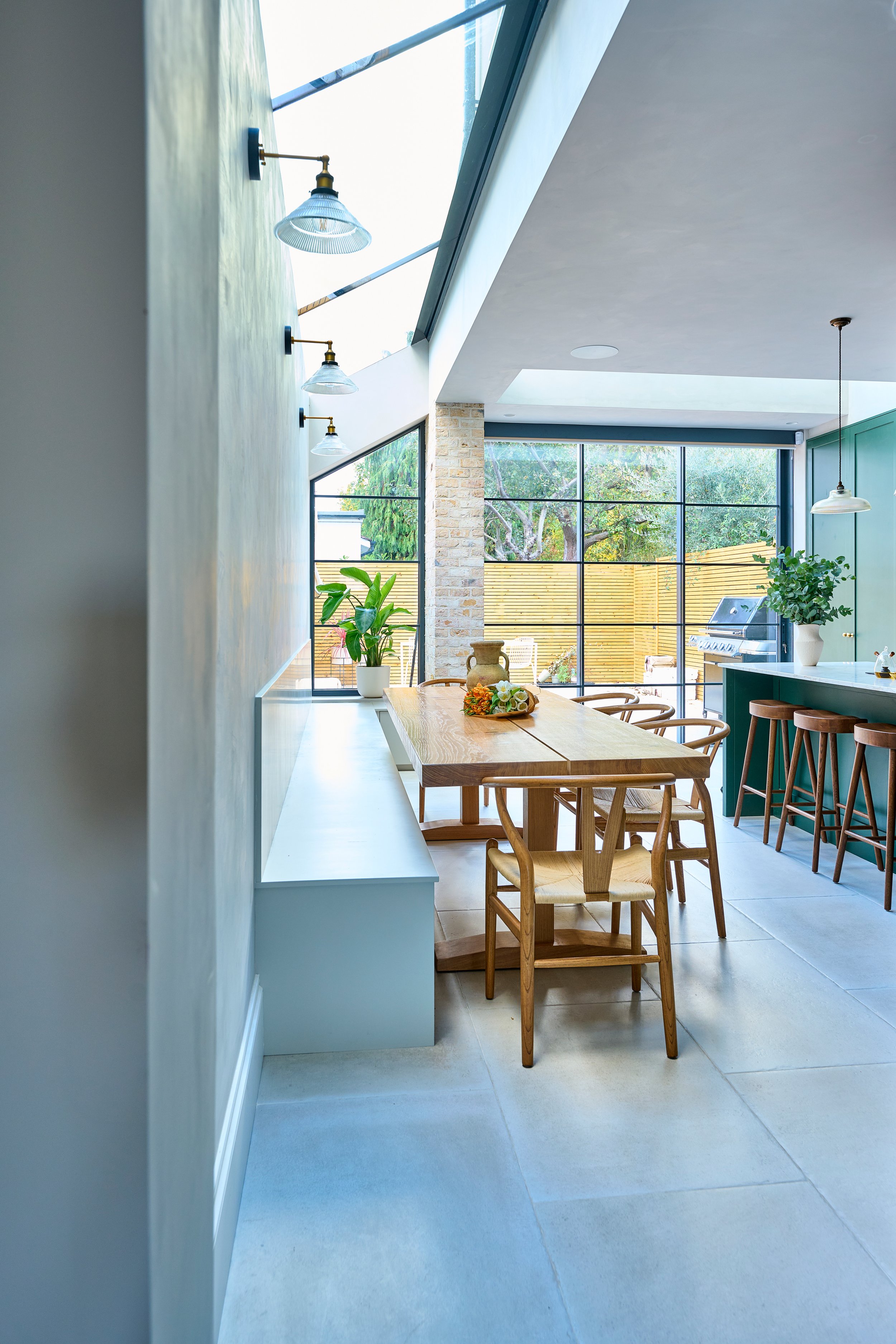 Modern dining area with wooden table and chairs, potted plants, large windows, and a grill outside.
