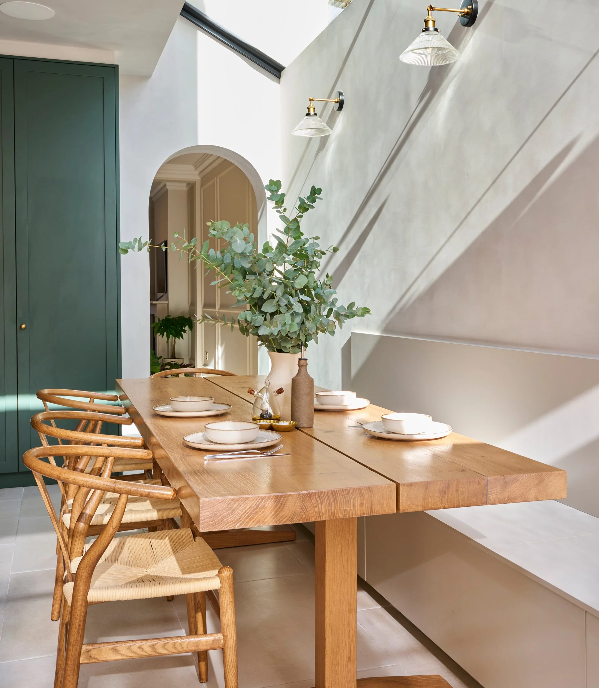 A modern dining area with a long wooden table set with white bowls, plates, and silverware. There are wooden chairs with woven seats around the table. A large green cabinet is in the background next to an arched doorway, and a plant with large green 