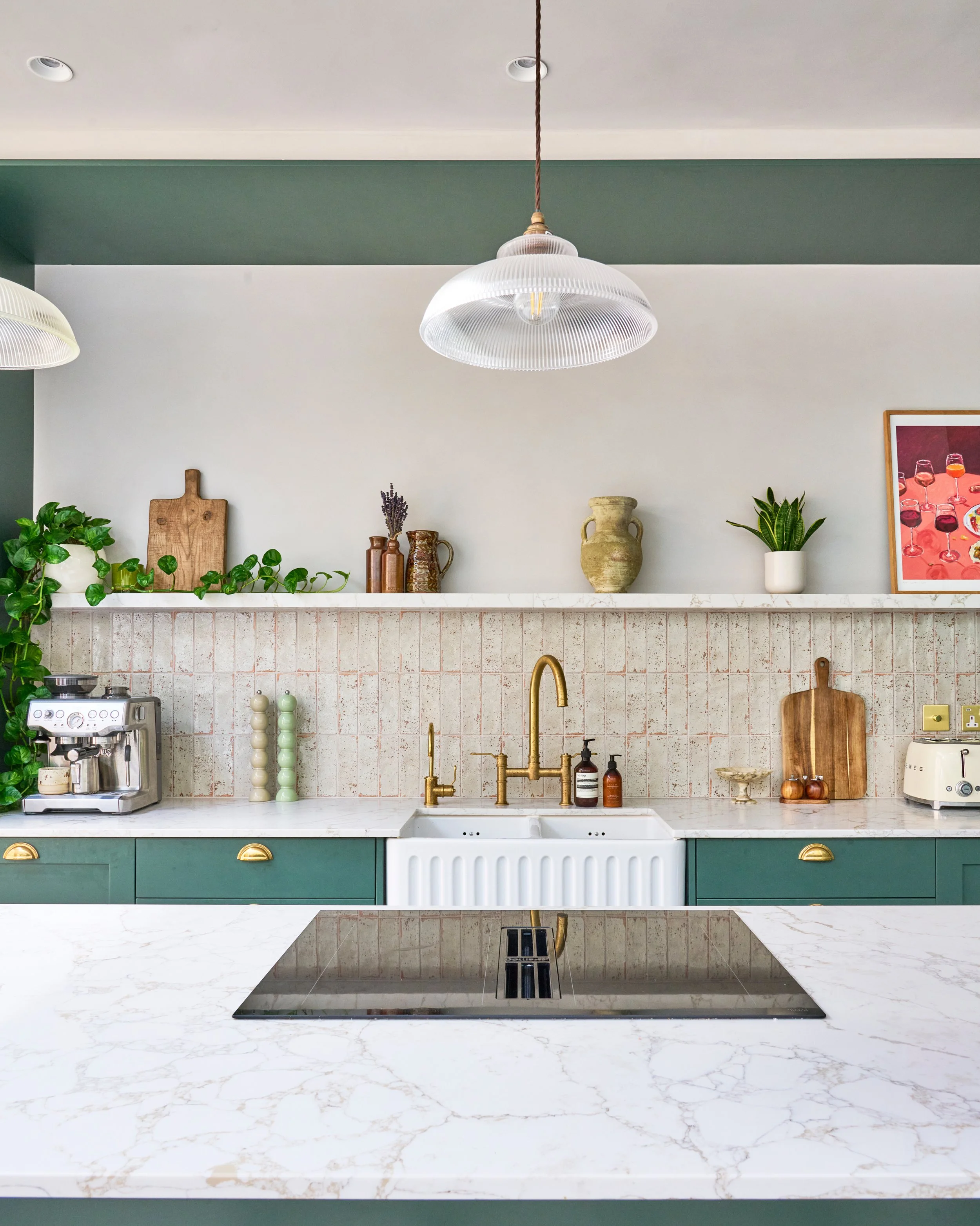Kitchen with marble countertops, gold faucet, hanging pendant lights, potted plants, decorative vases, cutting boards, framed artwork, espresso machine, and cooktop.