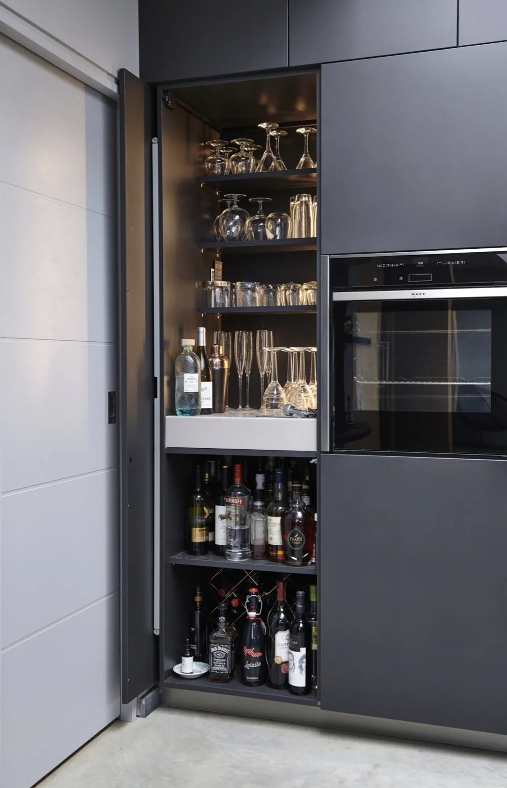 Open cabinet filled with various glassware, bottles of alcohol, and bar accessories, situated in a modern kitchen.