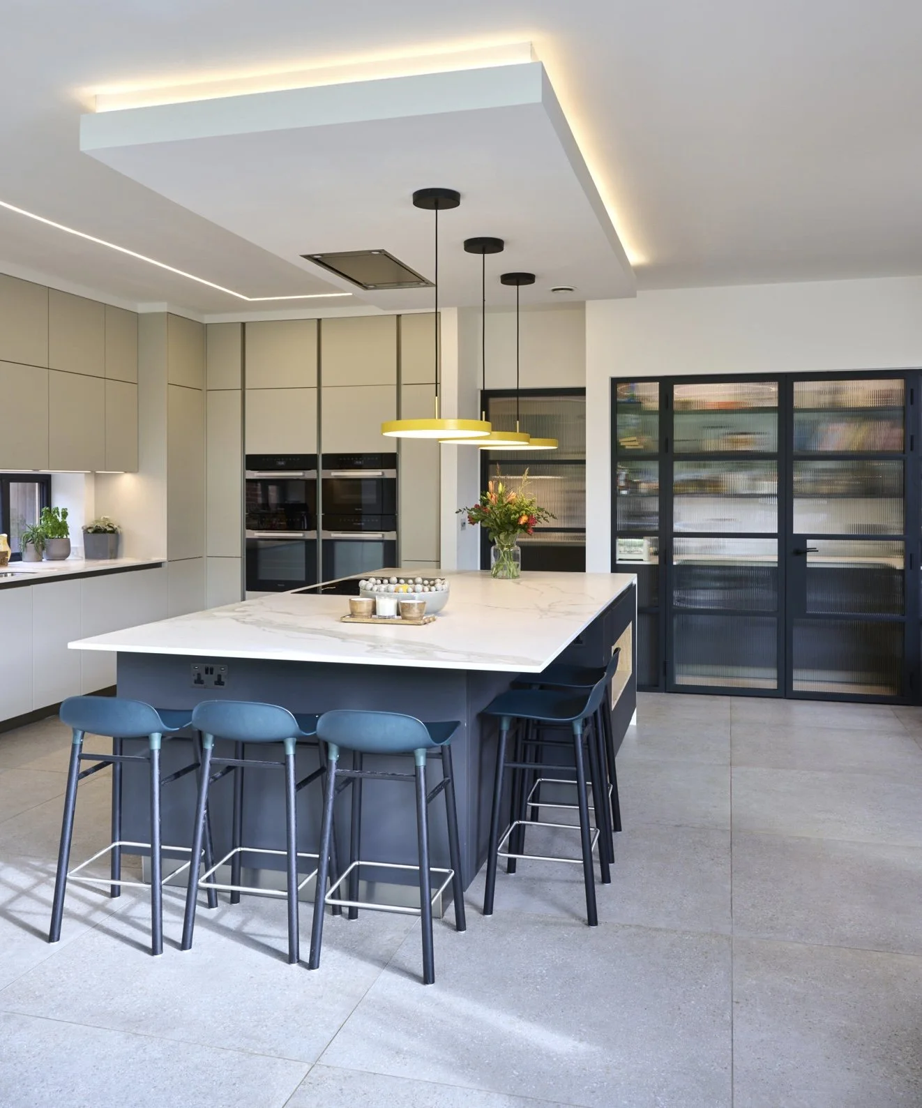 Modern kitchen with white marble island, yellow pendant lights, and black bar stools, with beige cabinets and sliding door.