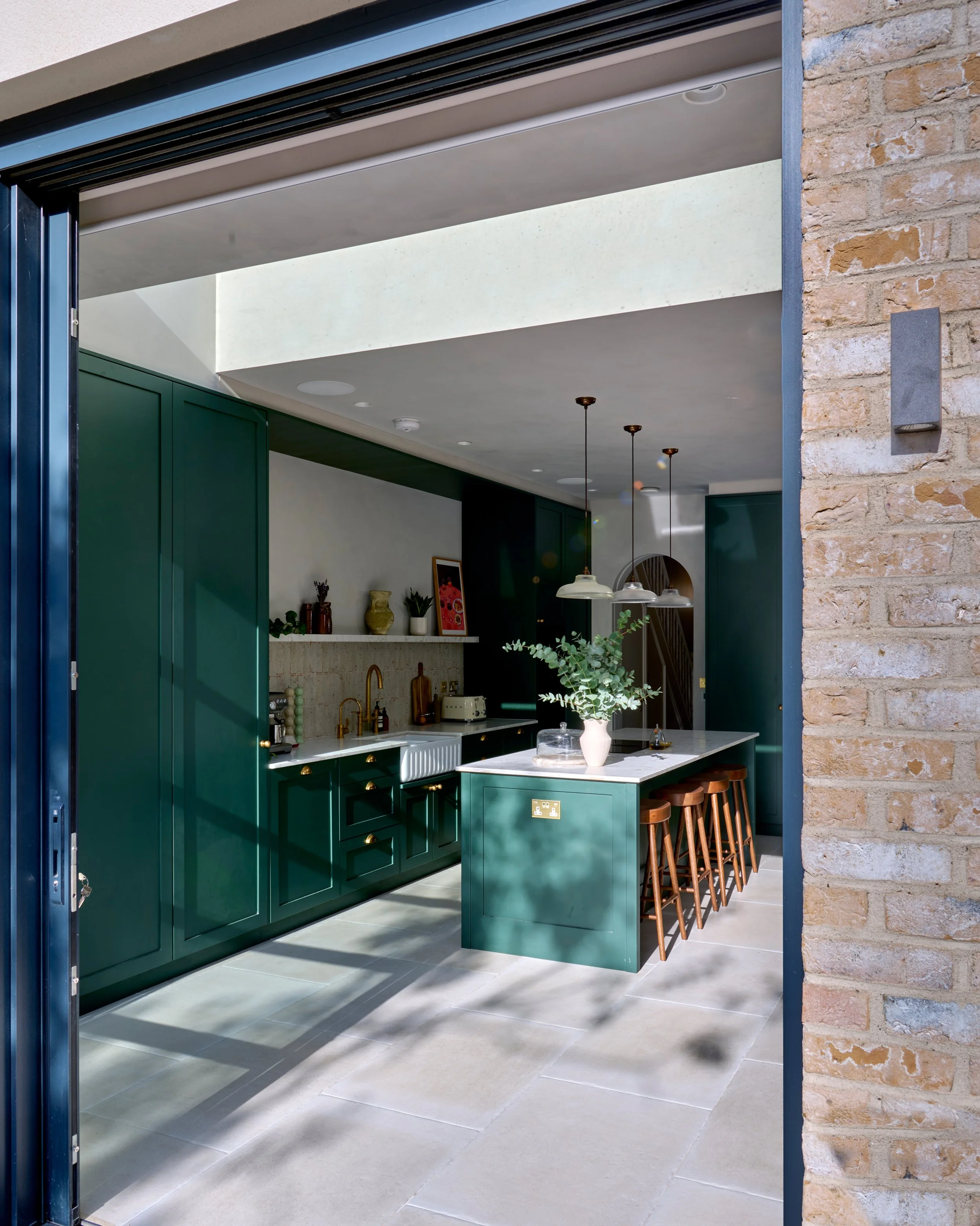 A modern kitchen with green cabinets, a white countertop with a vase of eucalyptus, hanging pendant lights, and wooden bar stools.