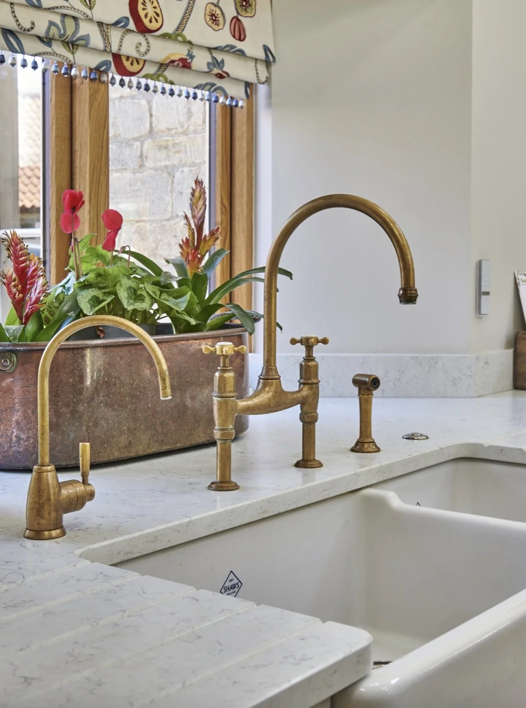 A kitchen sink with brass faucets, a flower planter with red and pink flowers, a window with a patterned valance, and a stone wall outside.