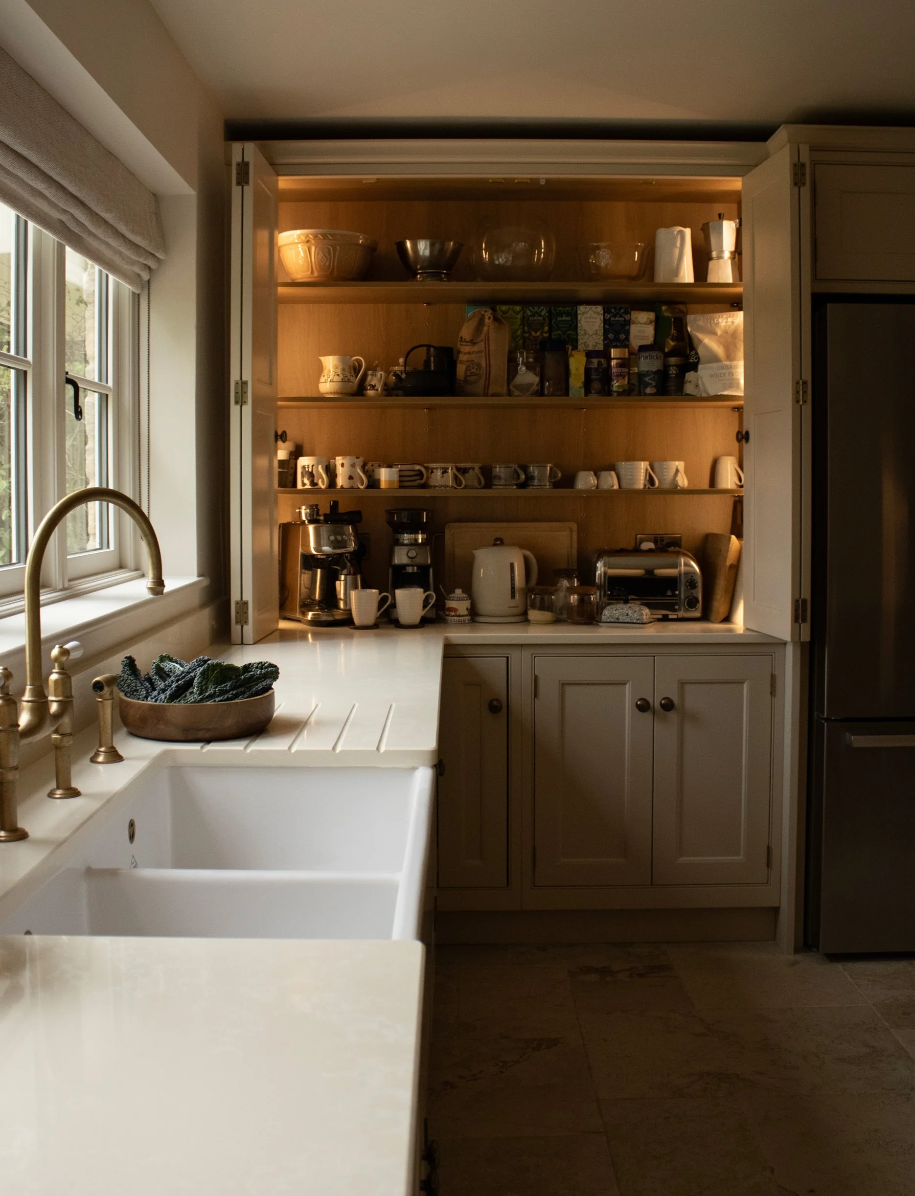 Kitchen cabinet with open doors, containing bowls, mugs, and kitchen appliances, with a window and a kitchen sink in the foreground.