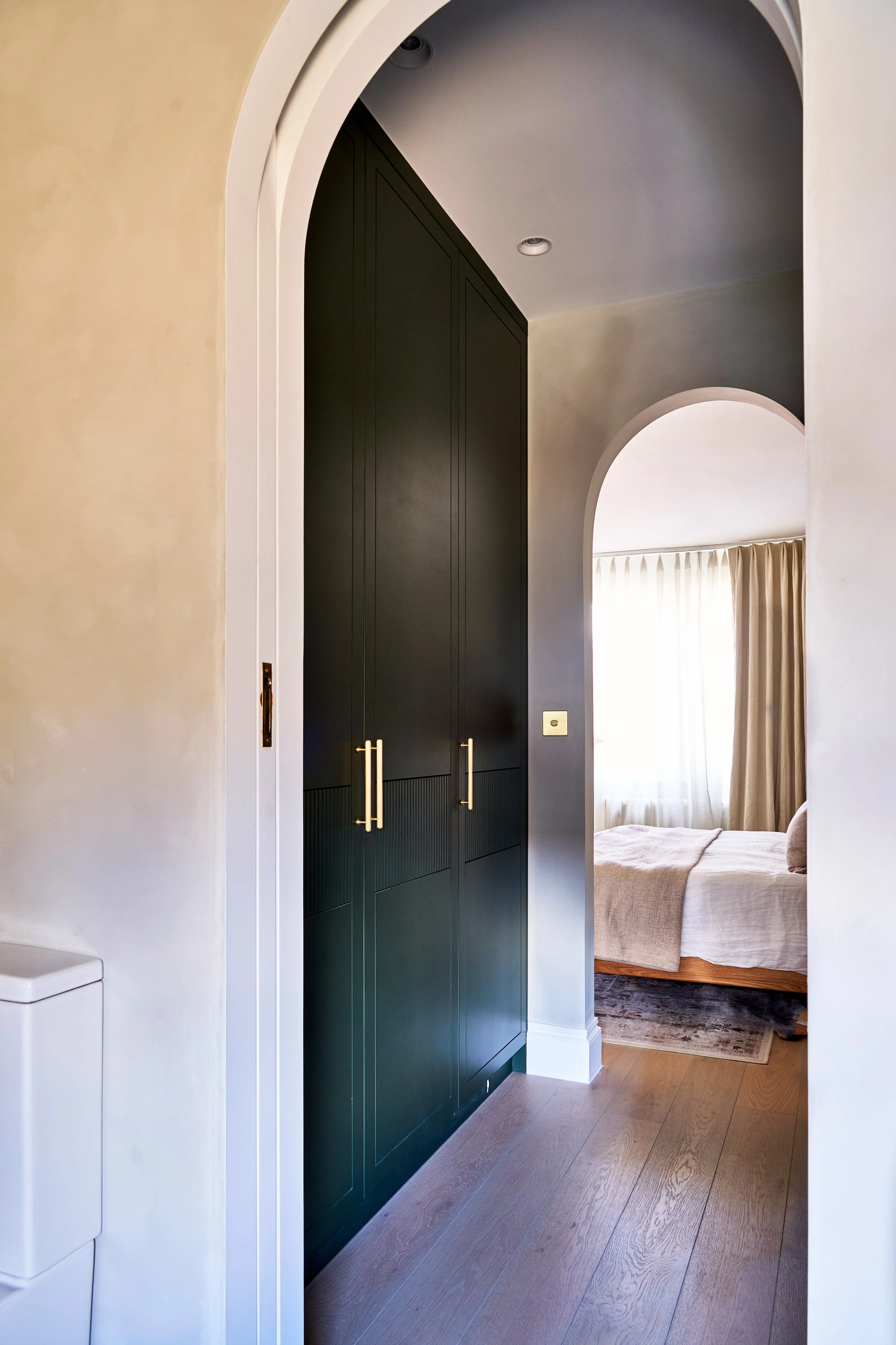 Interior view of a bedroom through an arched doorway, featuring a dark green wardrobe with gold handles on the left and a bed with beige bedding on the right, illuminated by natural light from a window with light curtains.