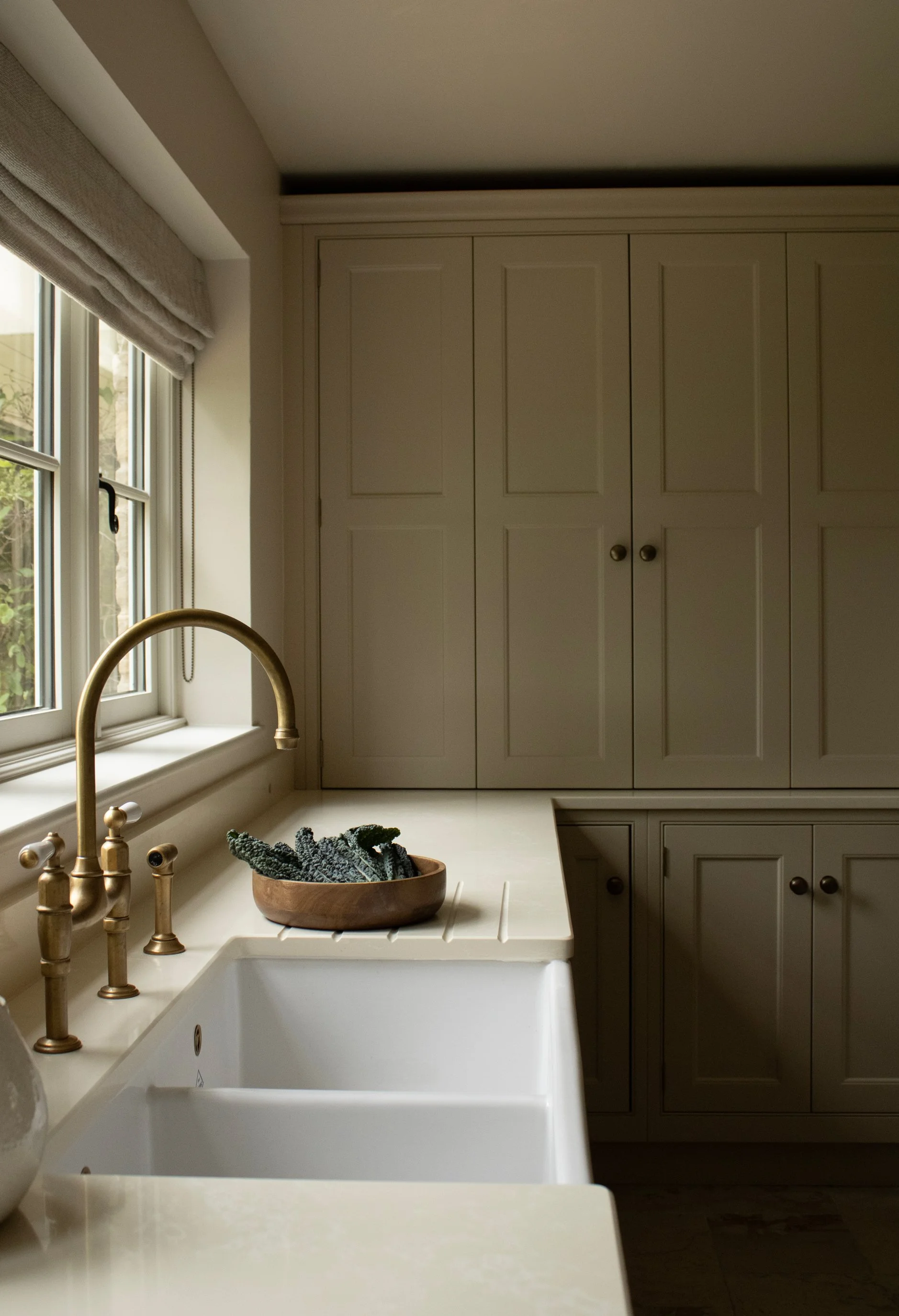 A kitchen with a white farmhouse sink, a brass faucet, a window with a beige roman shade, white cabinets with black knobs, and a wooden bowl with dark gray cloth or towels inside.