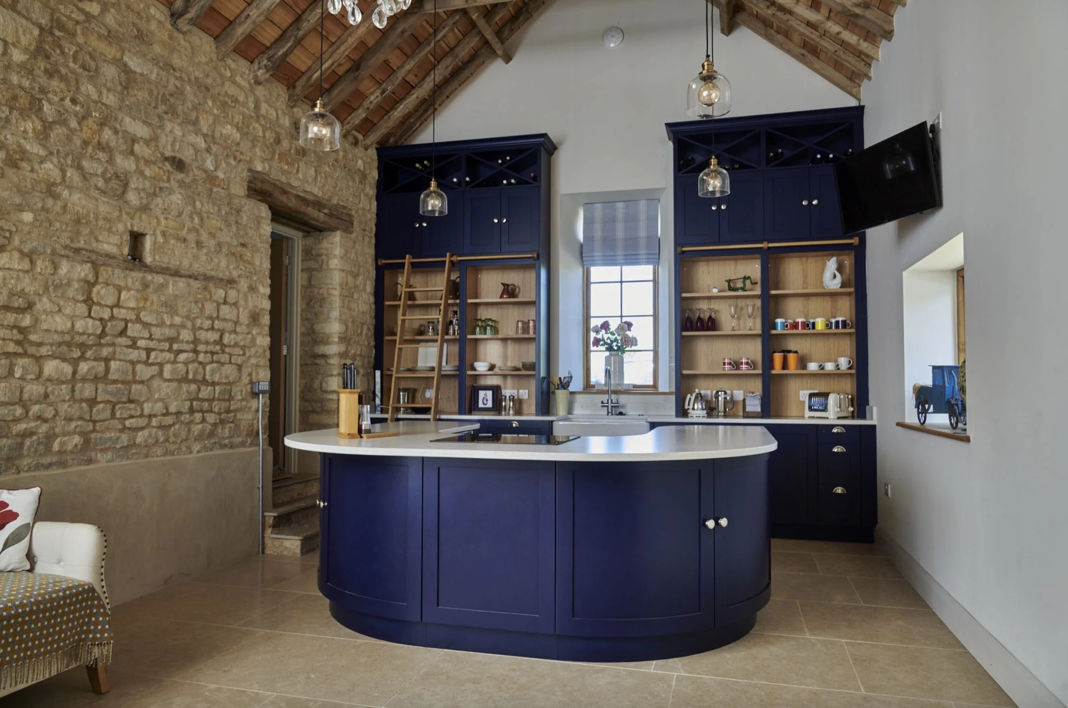 Kitchen with navy blue cabinets, open shelving, a central island with white countertop, a window with a checkered curtain, exposed brick and white walls, hanging glass pendant lights, and a small chalkboard on the wall.