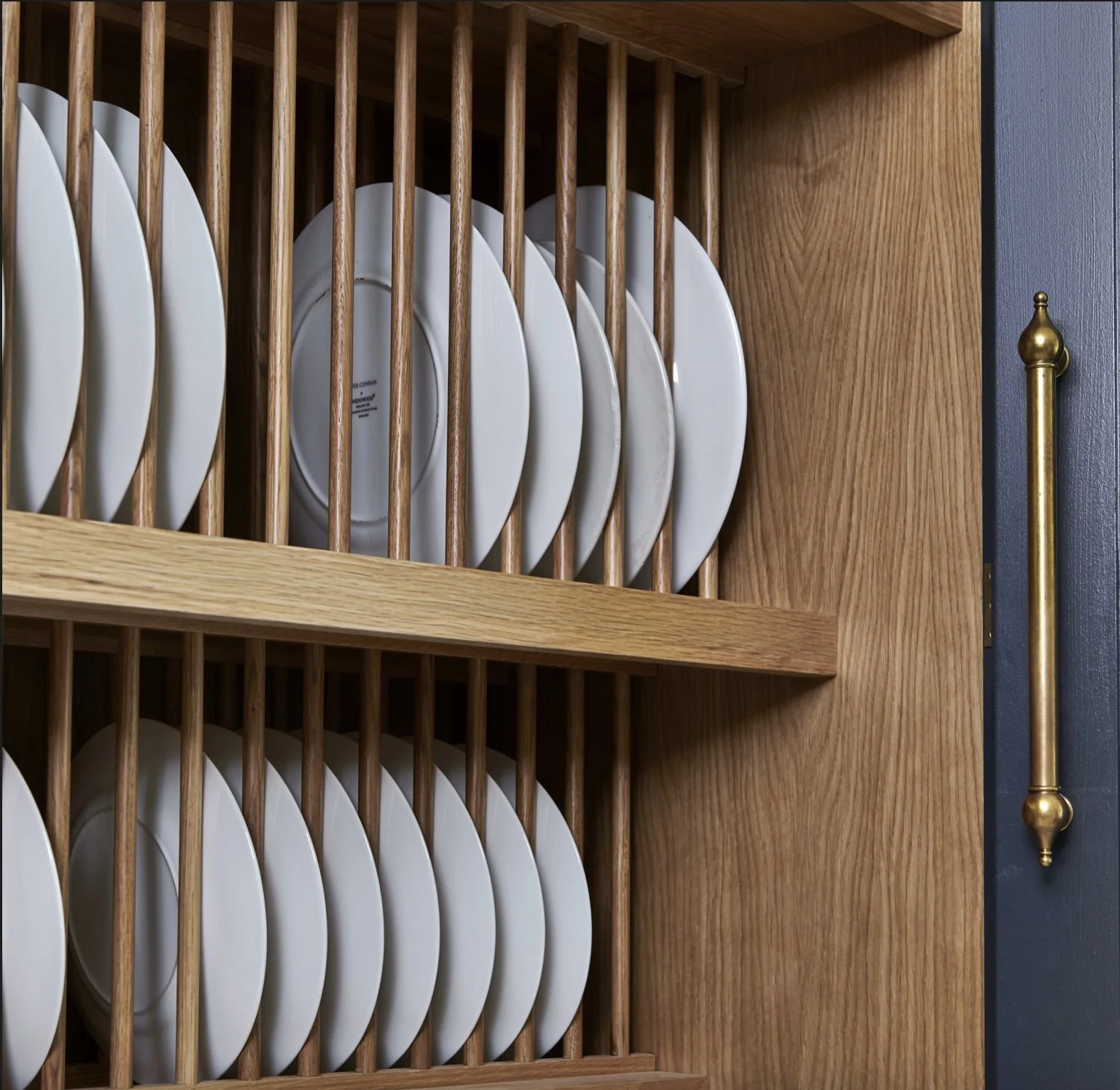 White plates stored vertically in a wooden dish rack with vertical slats, mounted inside a cabinet with a brass handle on the door.
