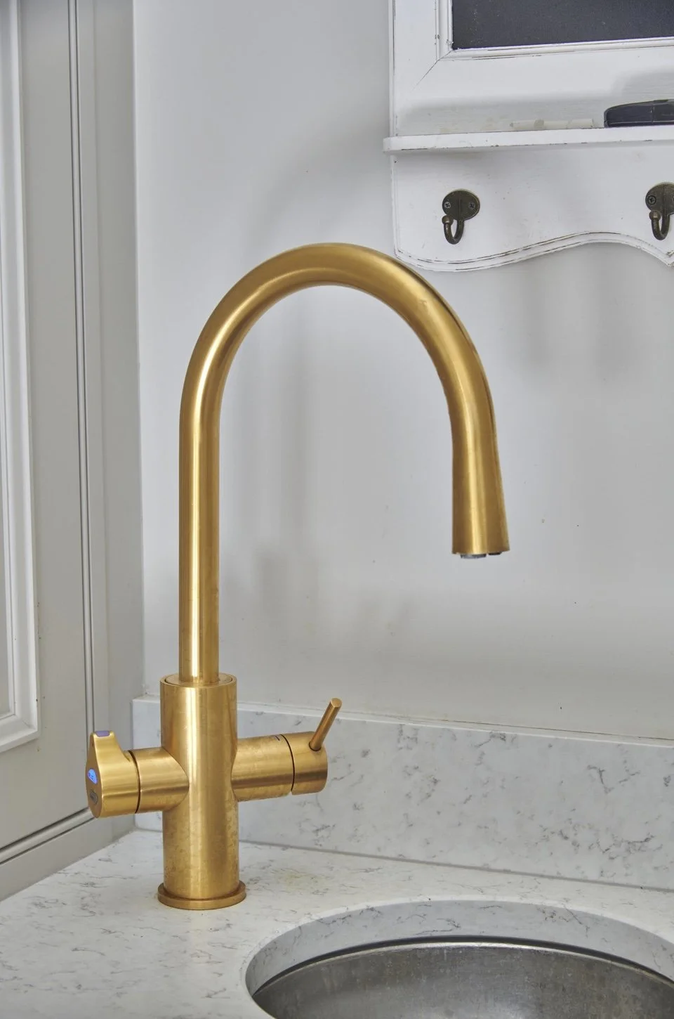 Close-up of a gold kitchen faucet mounted on a white marble countertop, with a white wall and cabinet above.