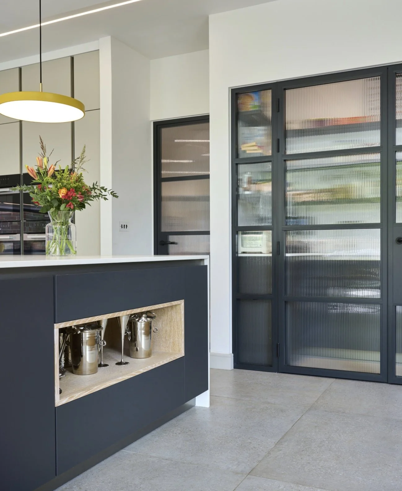 Modern kitchen with navy blue island, decorative open shelf with silver containers, glass vase with flowers, and textured glass sliding doors