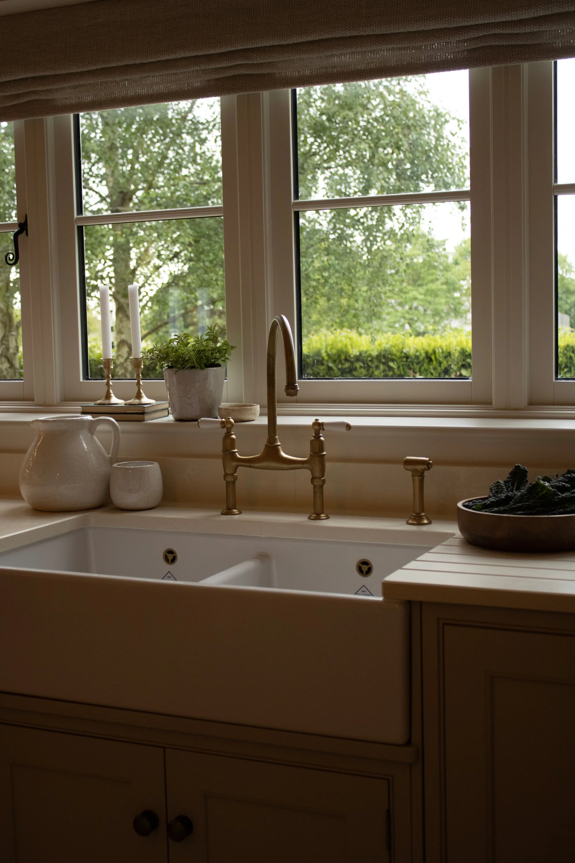 A kitchen window with a view of green trees outside. The windowsill has white candles, a potted plant, and a book. The kitchen has a beige countertop and a gold-colored faucet in the sink area.