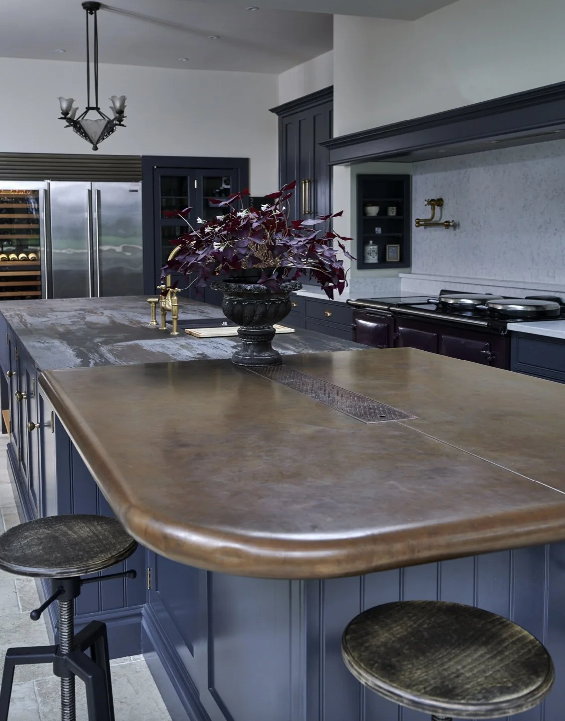A kitchen with a blue island, wooden countertop, and stools, decorated with a black vase holding purple leaves, stainless steel appliances, blue cabinets, and a hanging light fixture.