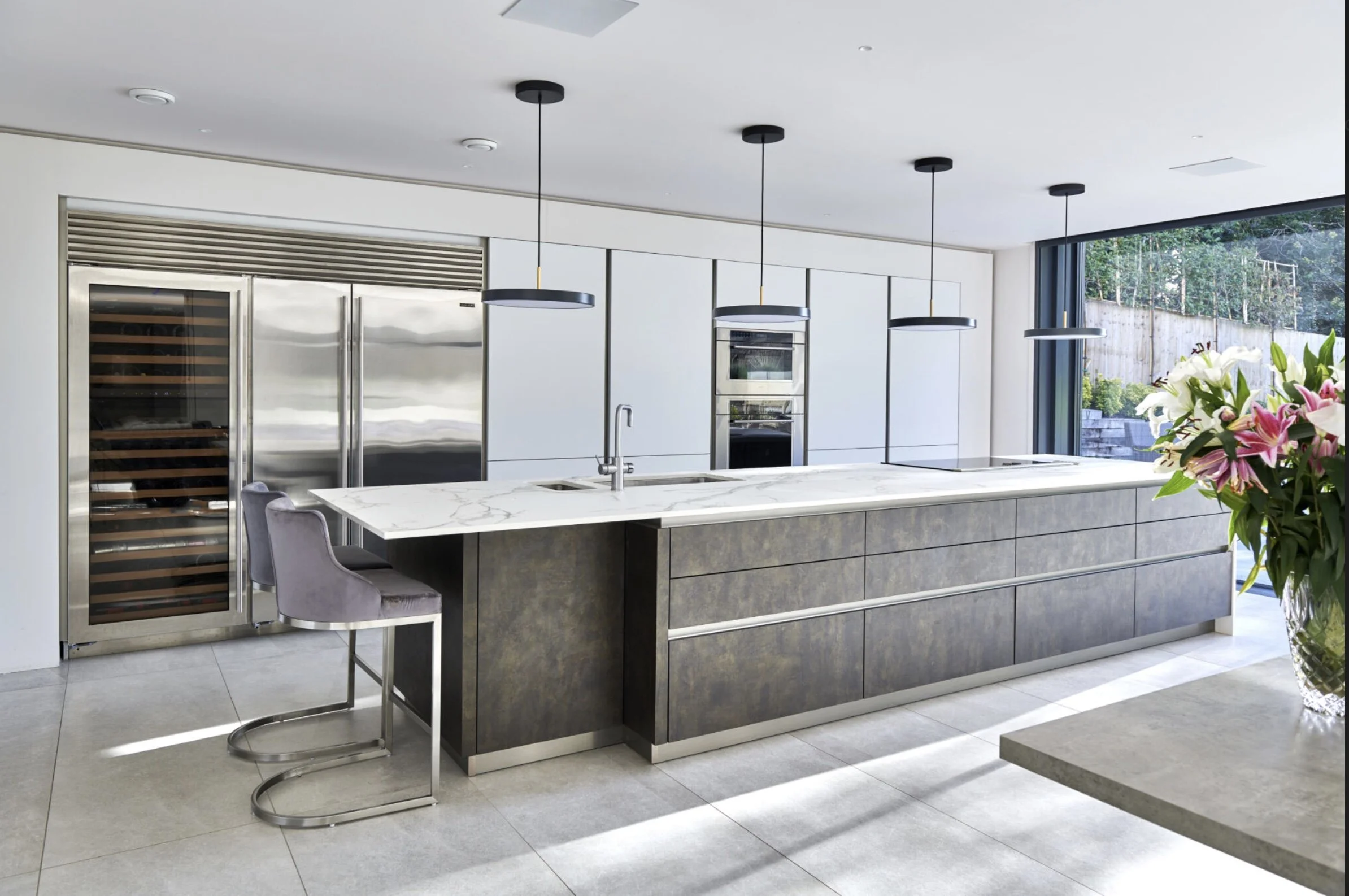 Modern kitchen with white walls, gray cabinets, marble island, stainless steel appliances, black pendant lights, large window with outdoor view, and a vase of pink and white lilies.