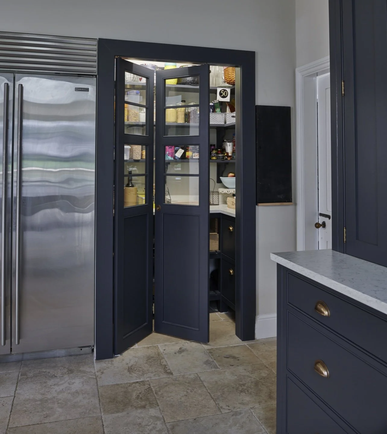 Open pantry cabinet revealing organized food storage in a kitchen with blue cabinets and stainless steel refrigerator.