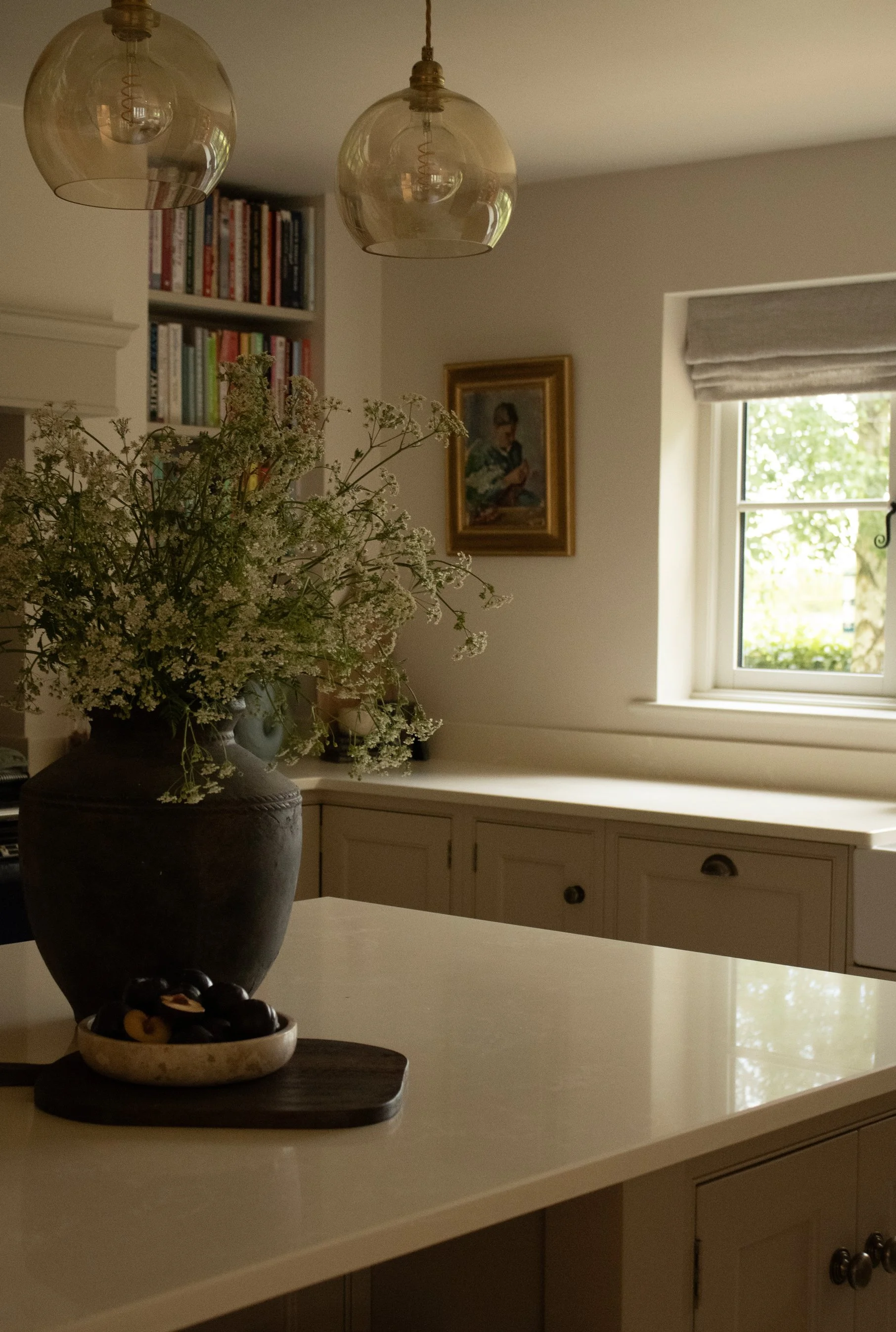 A kitchen corner with a white countertop, a large dark vase with white flowers, a small bowl of assorted olives and nuts, a window with a view of trees, a framed portrait on the wall, a bookshelf filled with books, and two hanging glass pendant light