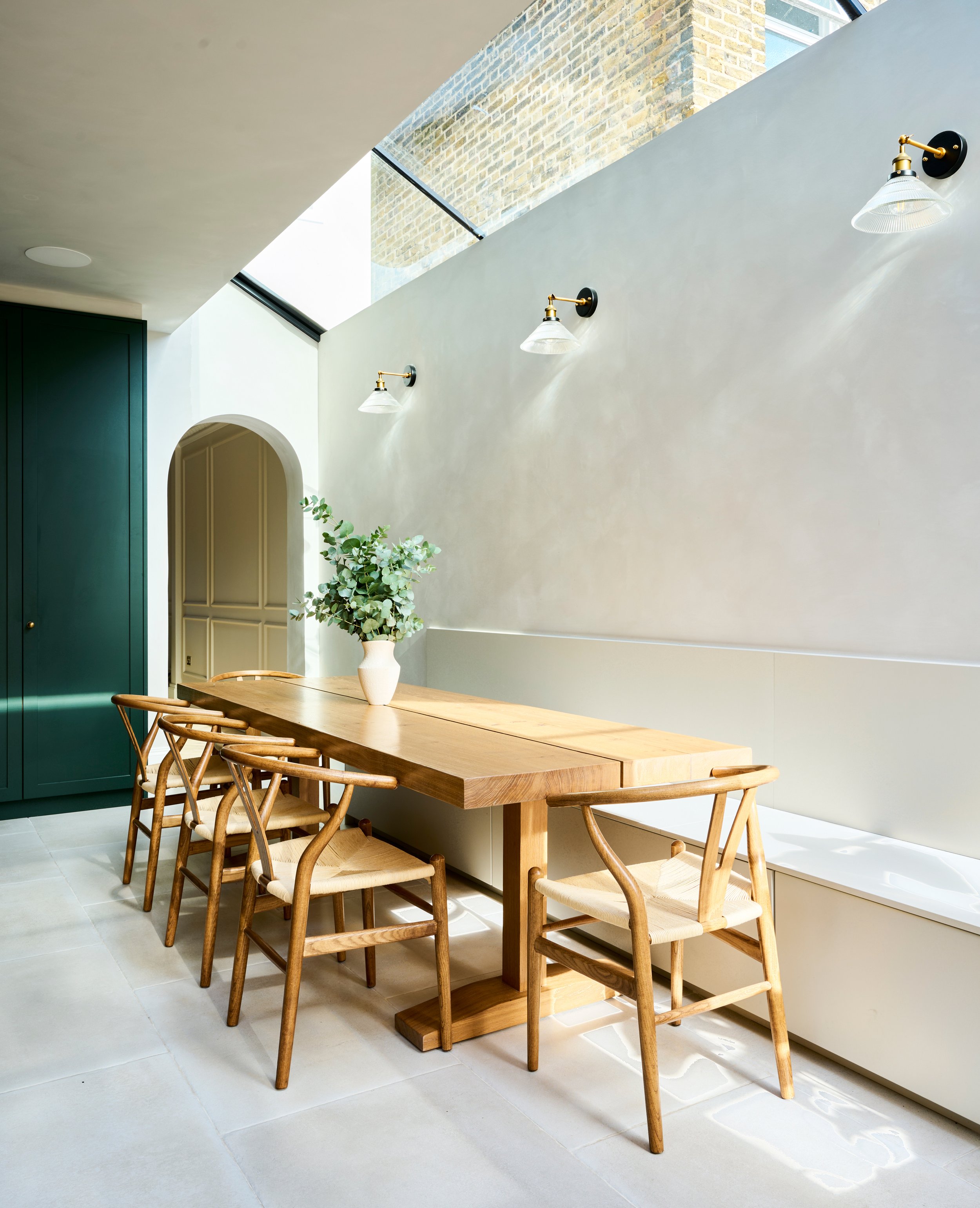 A modern dining area with a wooden table surrounded by eight matching wooden chairs with woven seats, a white vase with green eucalyptus leaves on the table, white tiled floor, a lime-green cabinet to the left, an arched doorway, a large plant, and w