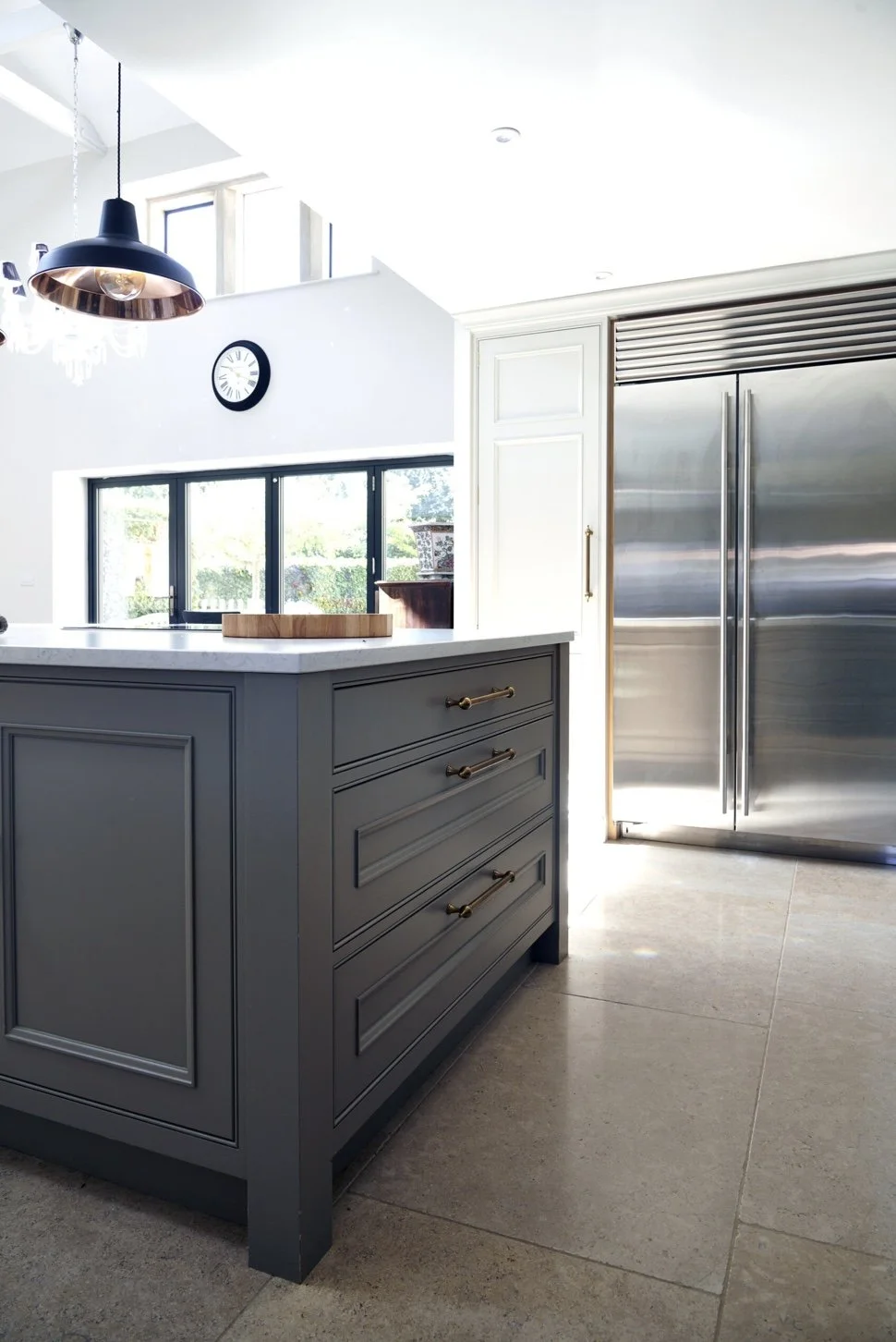 A modern kitchen interior with gray cabinetry, a white marble countertop, stainless steel refrigerator, black-framed sliding door windows, a wall clock, and hanging lights.