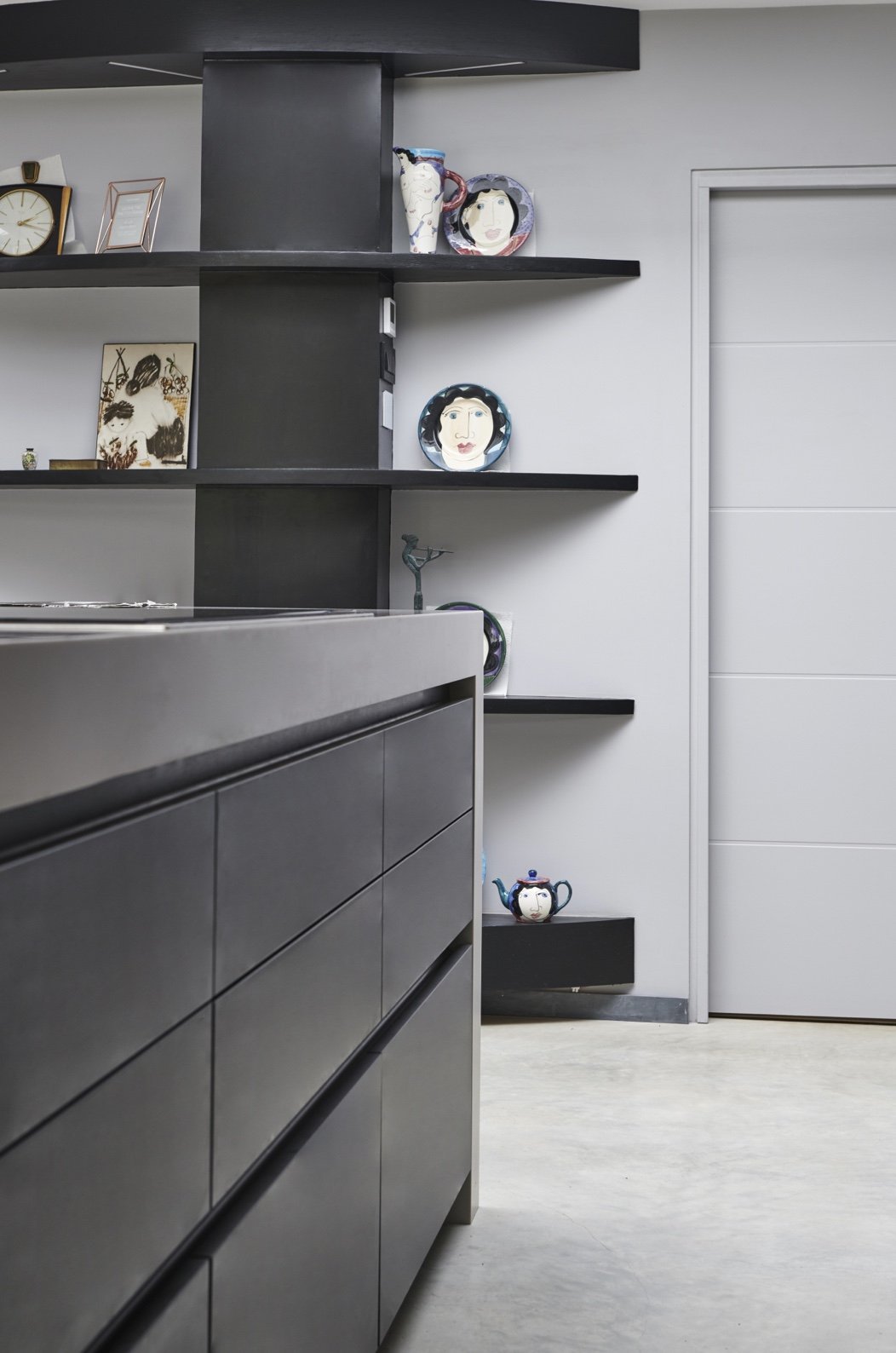 View of a modern kitchen with black and gray cabinets and decorative plates on the wall shelf, a white door, and a concrete floor.
