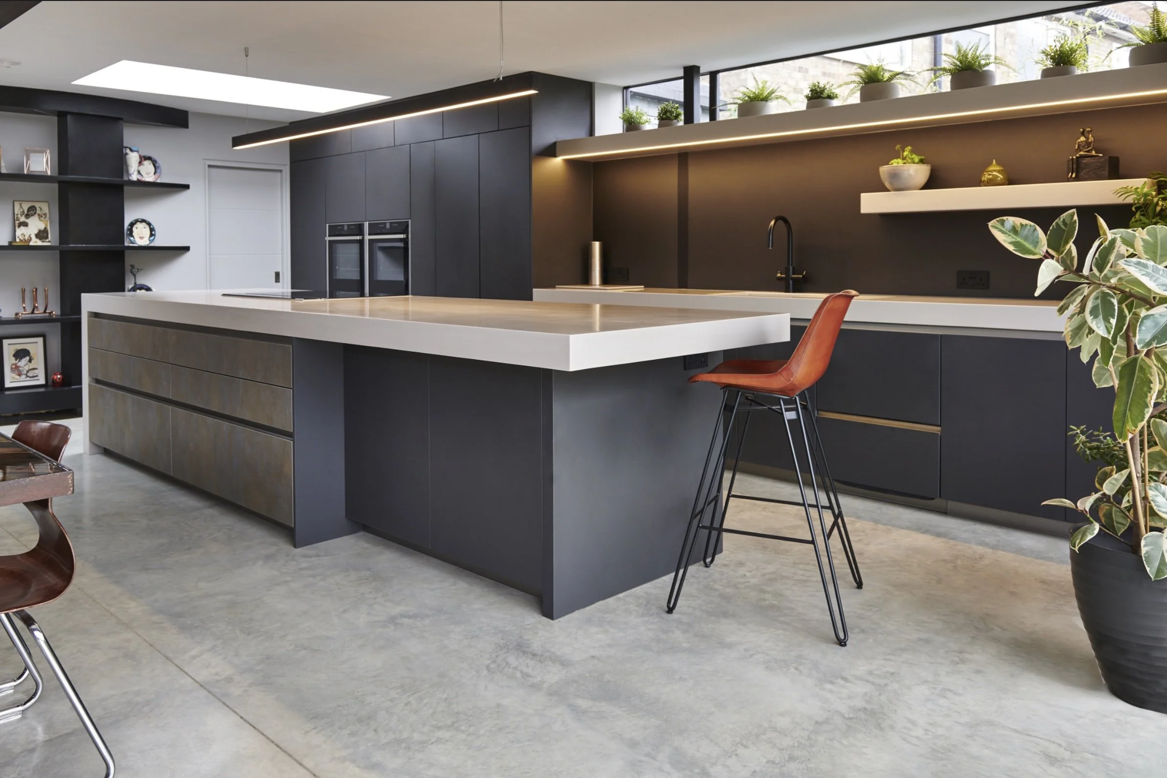 Modern kitchen with dark cabinets, white countertops, open shelving with decorative items, potted plants, and a kitchen island with a barstool.
