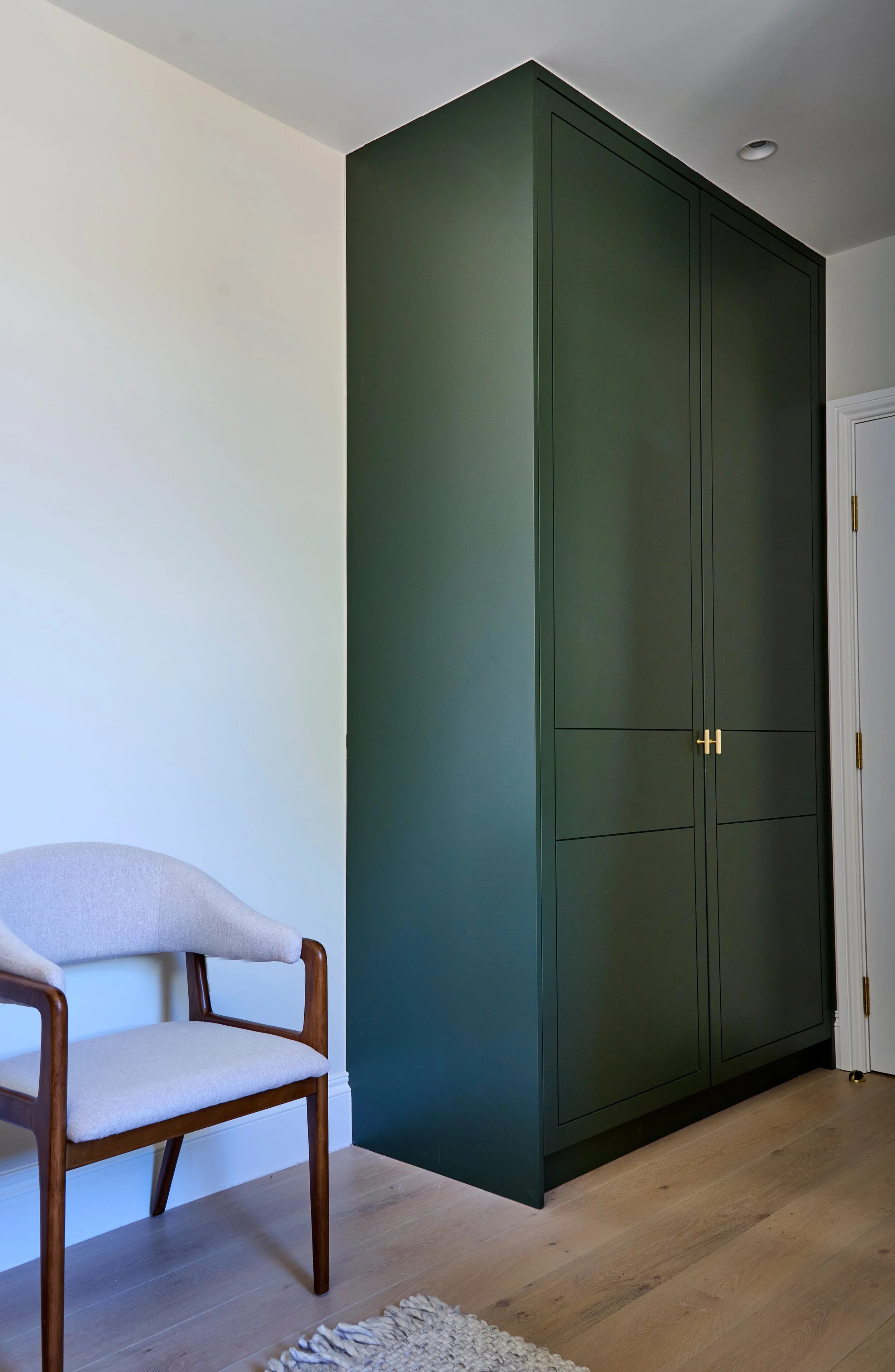 A corner of a room with two wooden chairs with beige cushions, a large dark green cabinet, a white wall, a hardwood floor, and a rug.