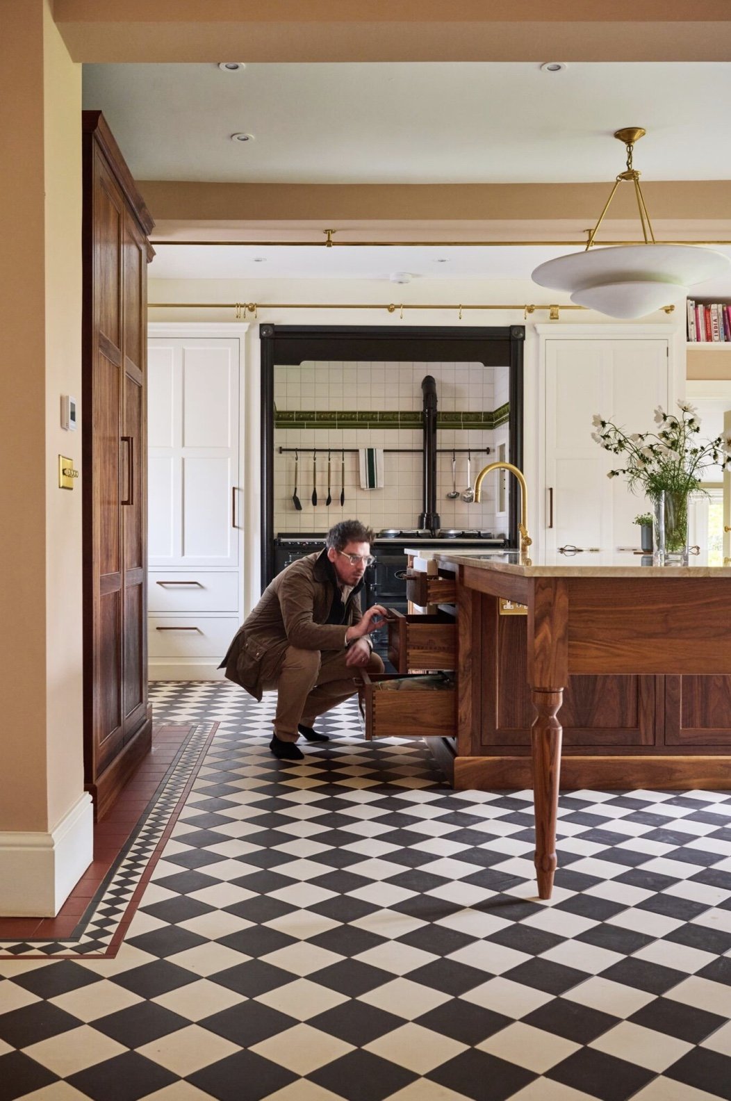 A man squatting down and opening a drawer in a wooden kitchen island in a spacious, well-lit kitchen with black and white checkered floor tiles, white cabinets, and a brass-colored pendant light.