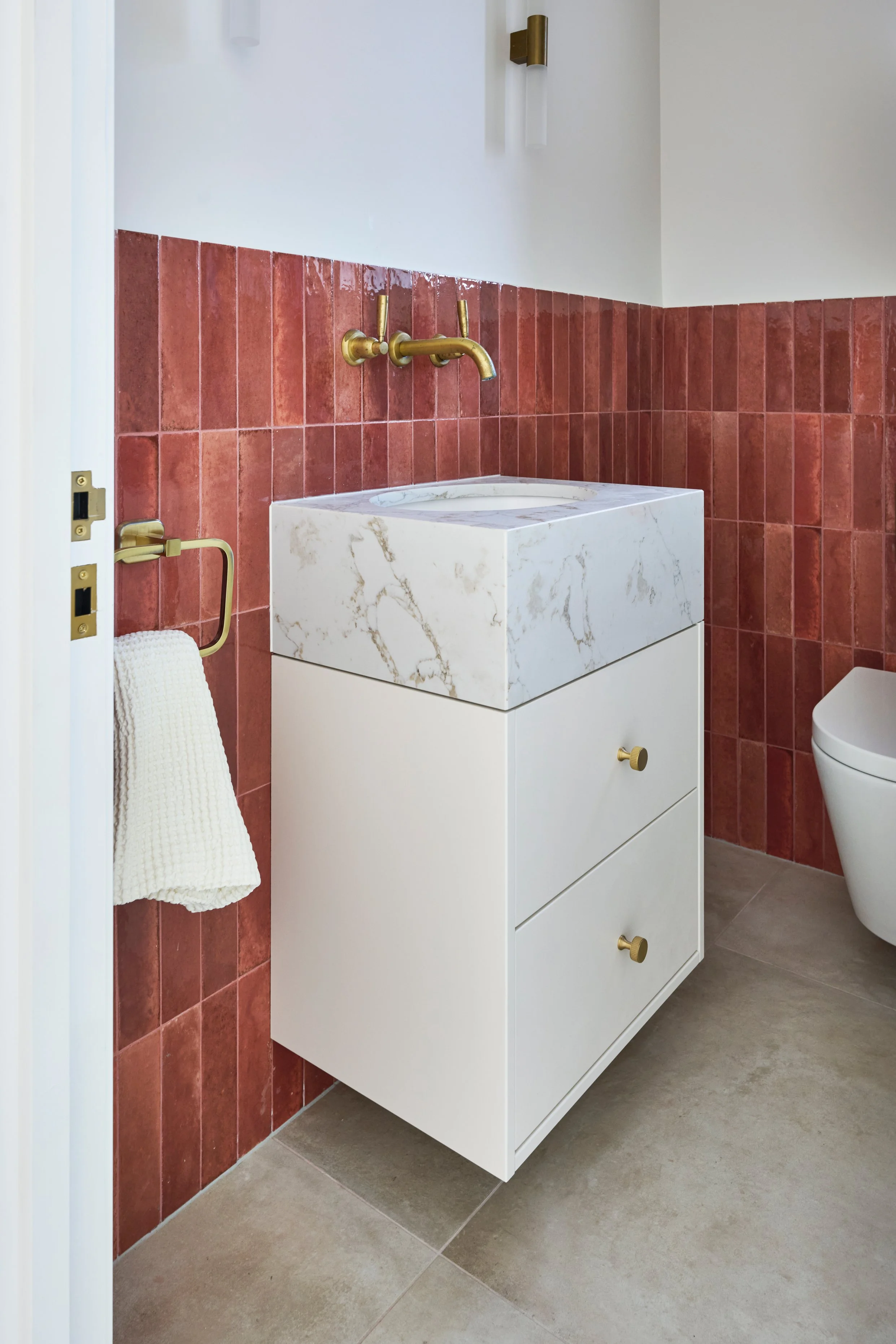 A modern bathroom featuring a white marble sink with gold fixtures, red tile wall, beige towel on a gold towel ring, and beige tiled floor.