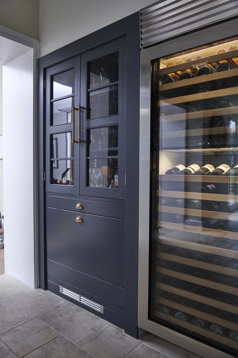 A dark blue cabinet with glass-paneled doors displaying glassware, positioned next to a wine cooler filled with wine bottles on wooden shelves.