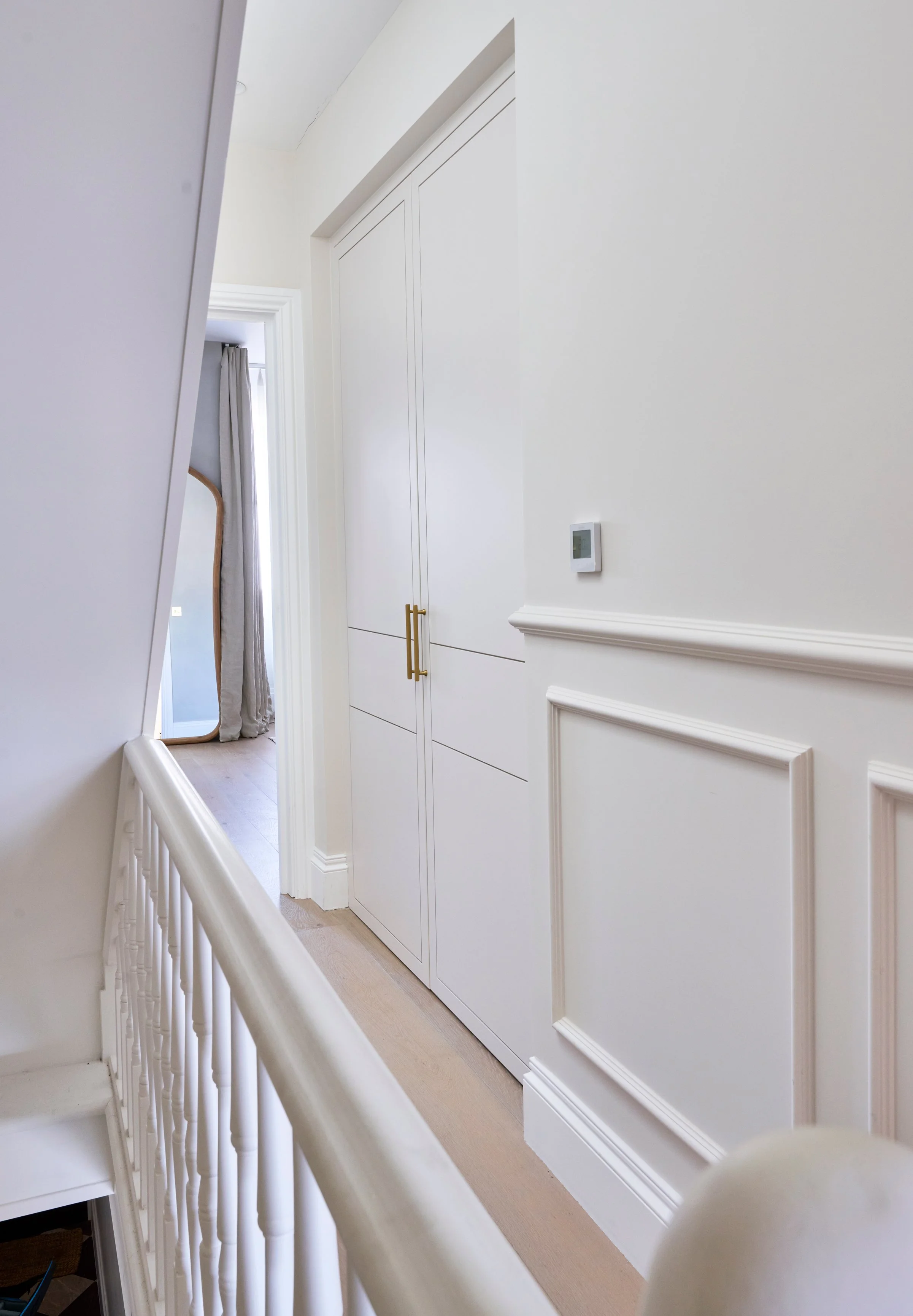 A bright, modern hallway with white walls, a thermostat on the wall, a white door with gold handles, and a hallway leading into another room with a window and curtain, partially visible through an open door.