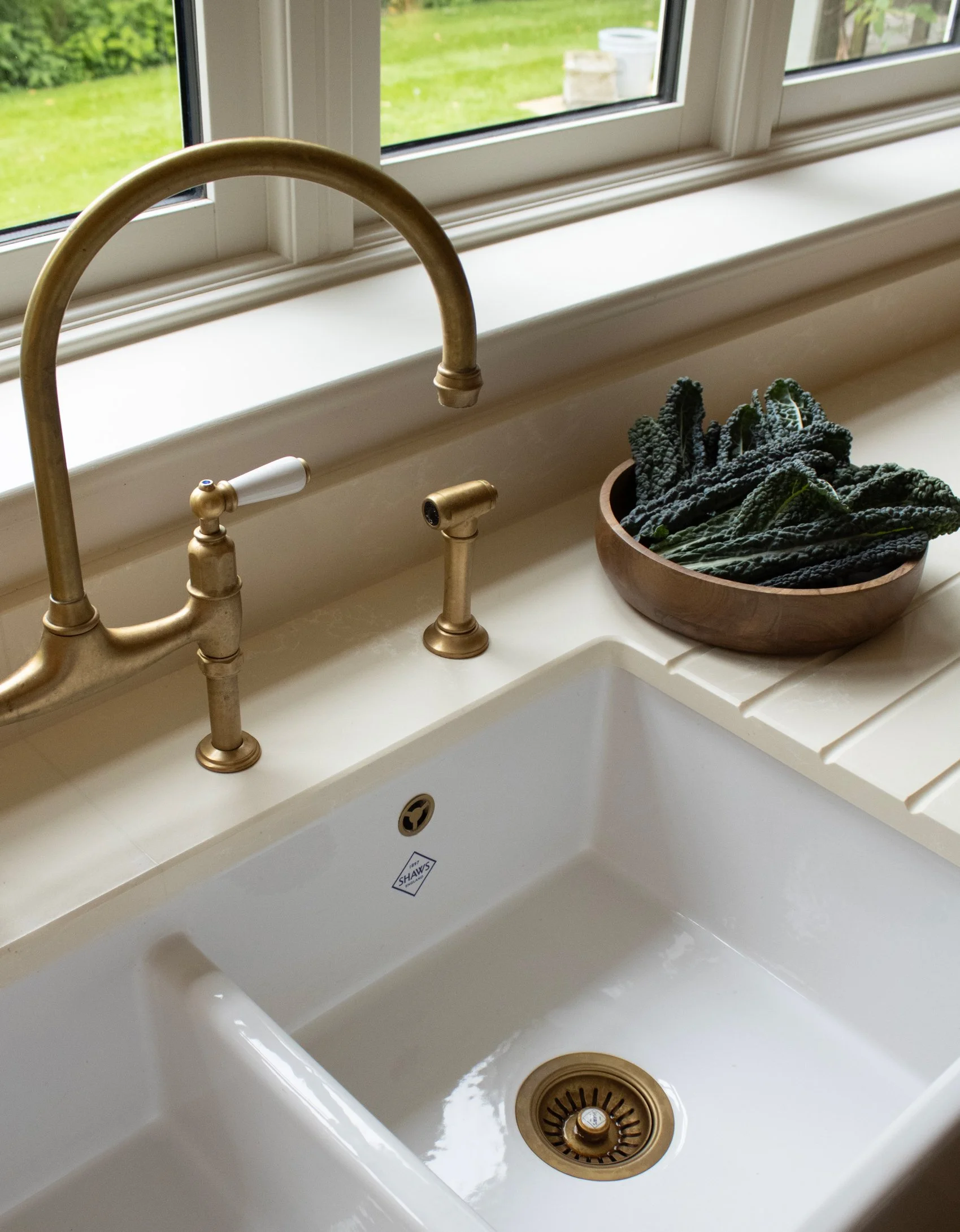Kitchen sink with a brass faucet and a wooden bowl containing dark leafy greens on the right side, outdoor view through a window in the background.